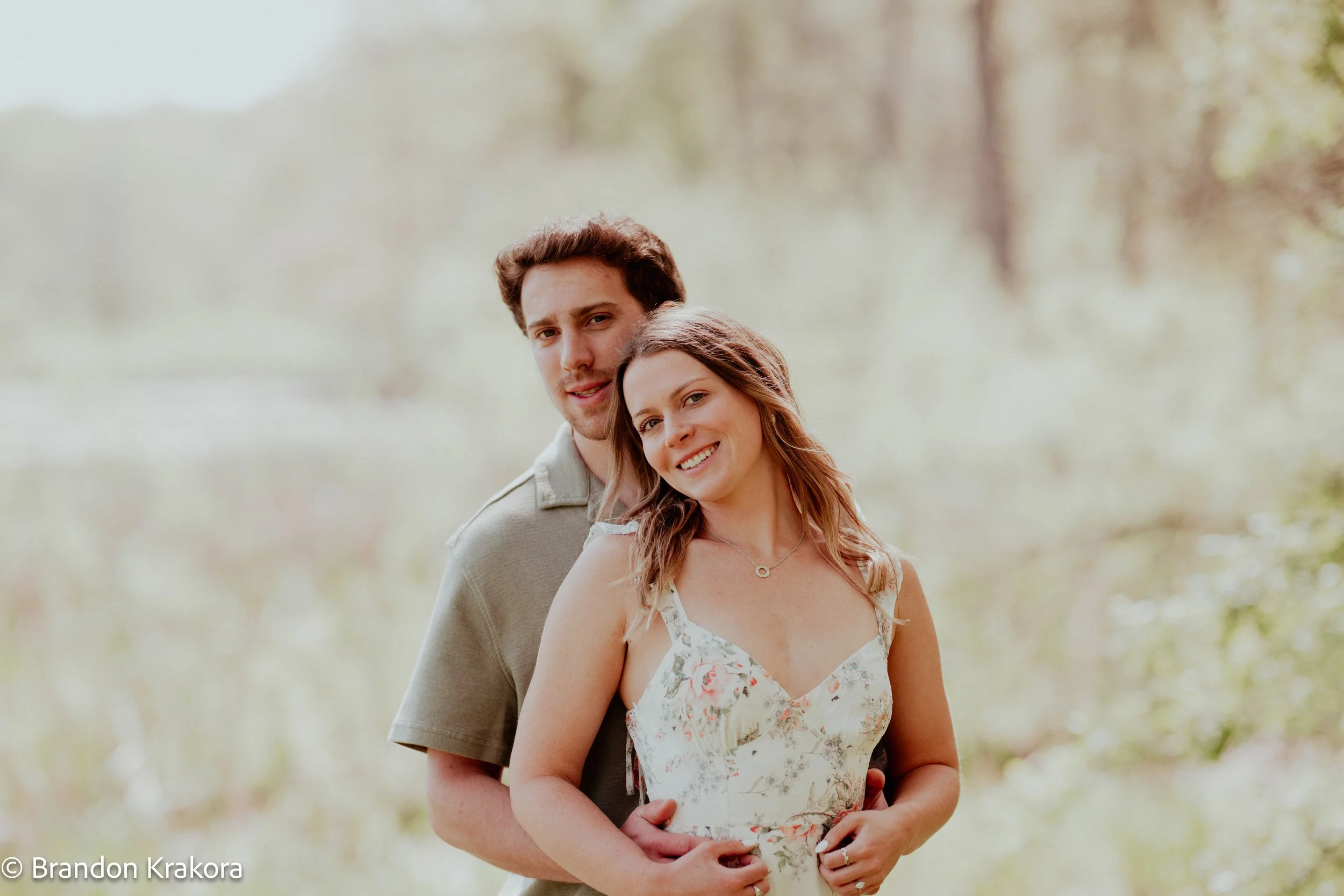 A young couple standing closely together outdoors, smiling, with a blurred nature background.