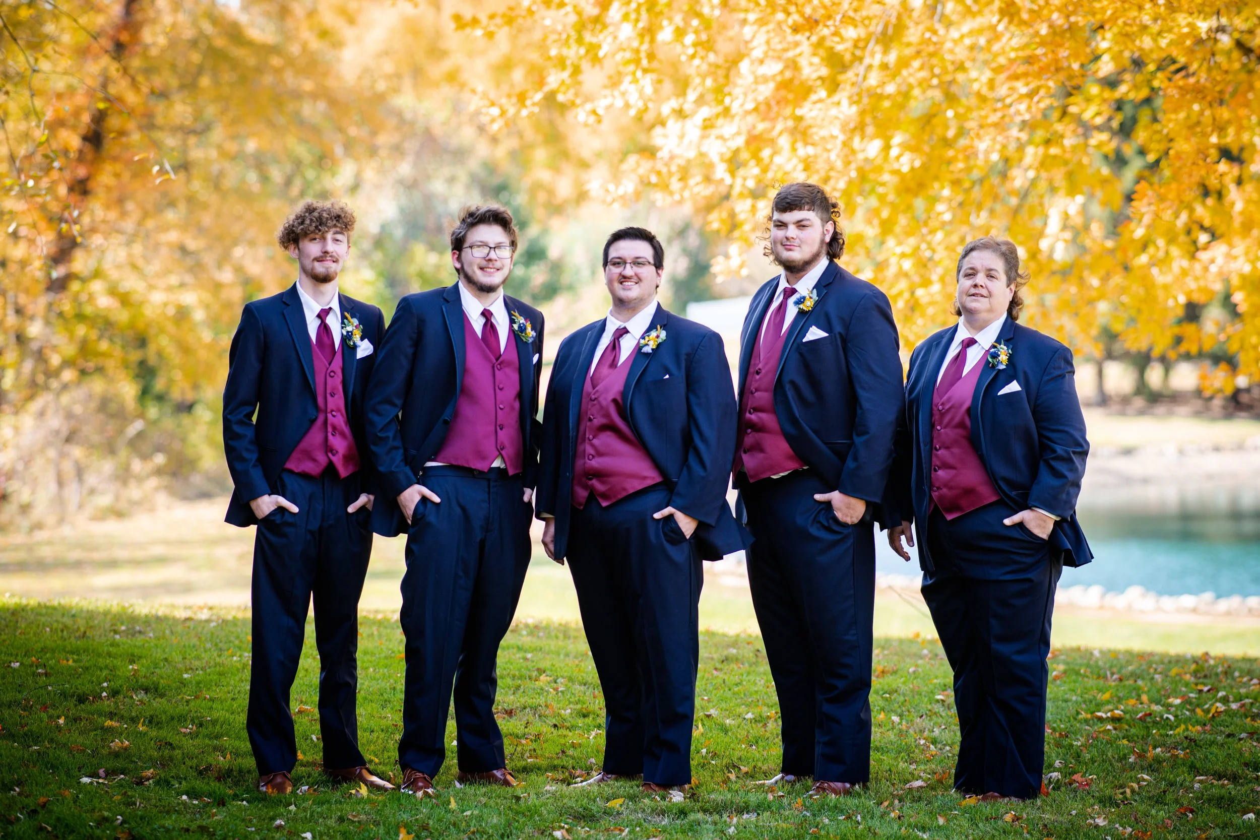 Group of six men dressed in formal suits with maroon vests and white shirts, standing outdoors on grass with autumn-colored trees in the background.