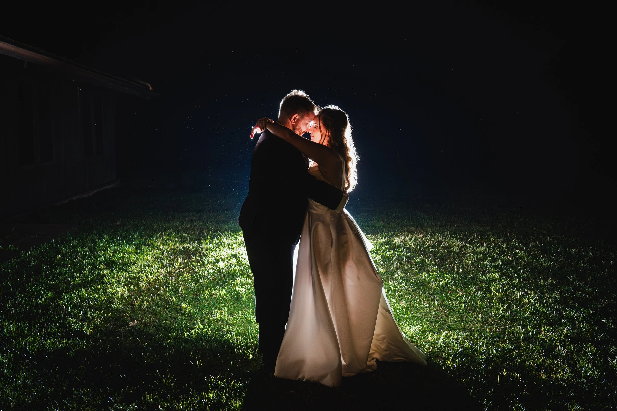 A couple in wedding attire dancing outdoors at night, illuminated from behind creating a halo effect around them, with dark background and grass underfoot.