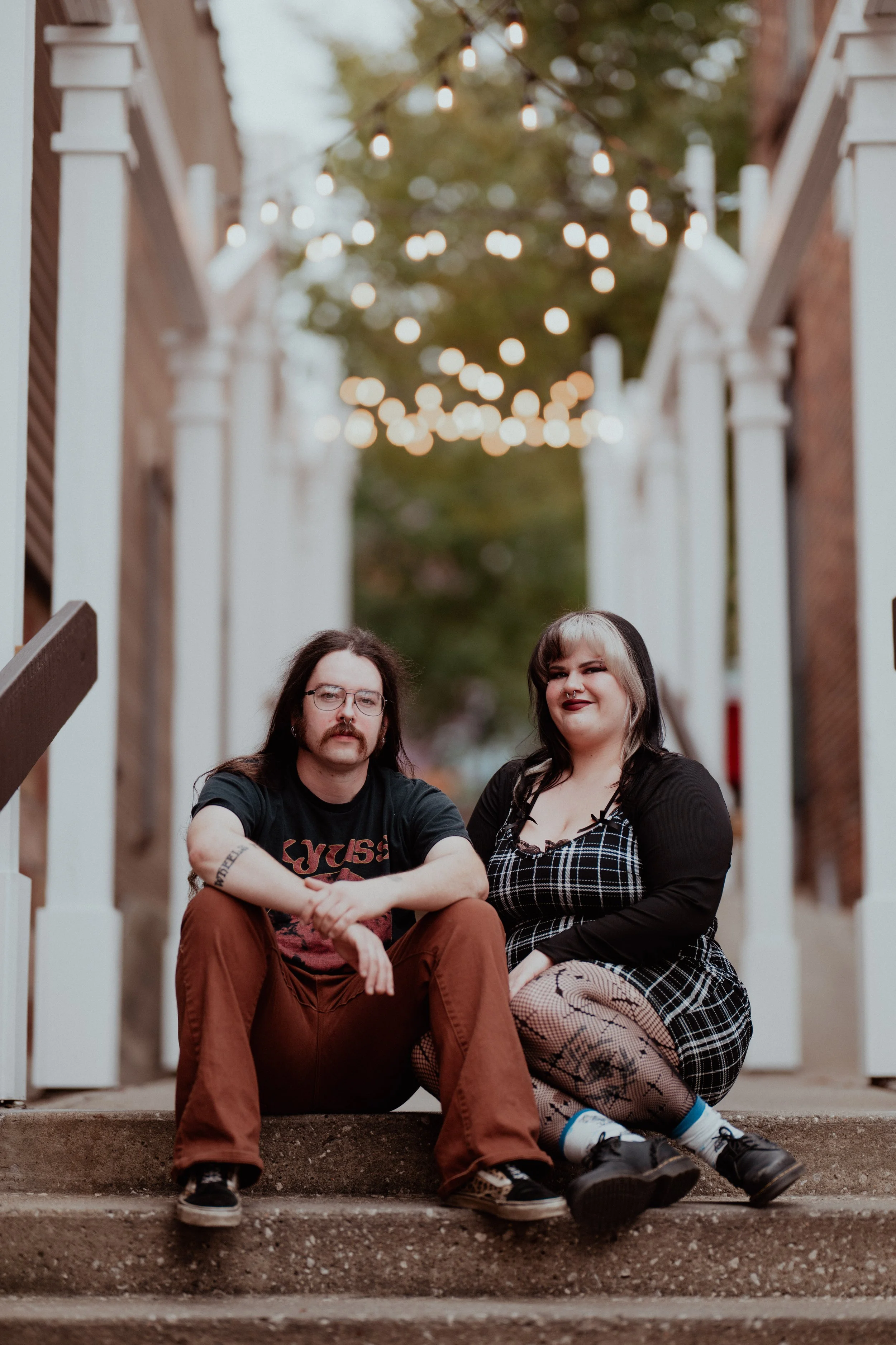 A man and woman sitting on steps outdoors with string lights hanging above them, standing between two brick buildings.