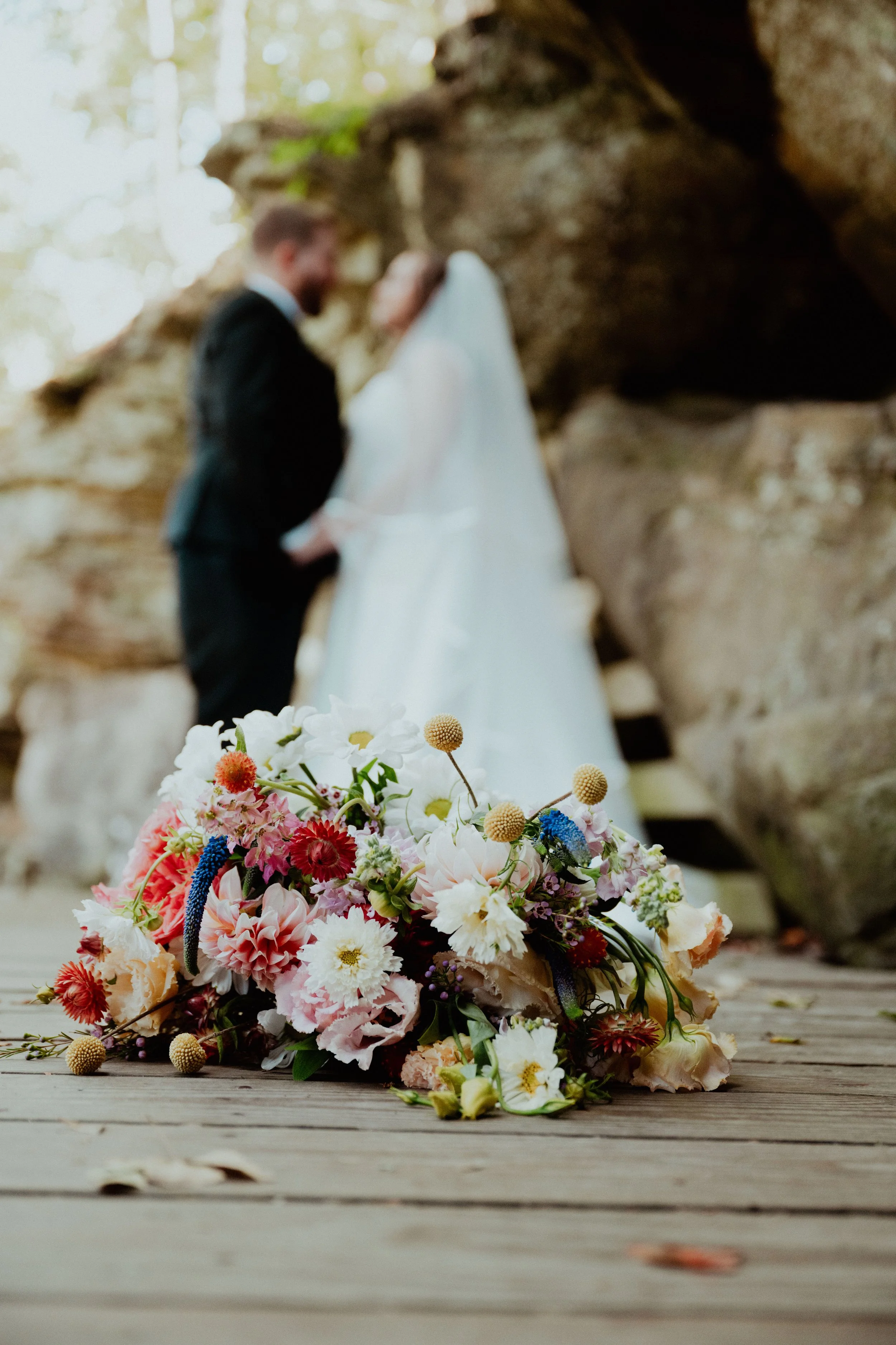 A colorful flower bouquet lies on a wooden surface in the foreground, with a blurred wedding couple in formal attire holding hands and standing near a rocky formation in the background.