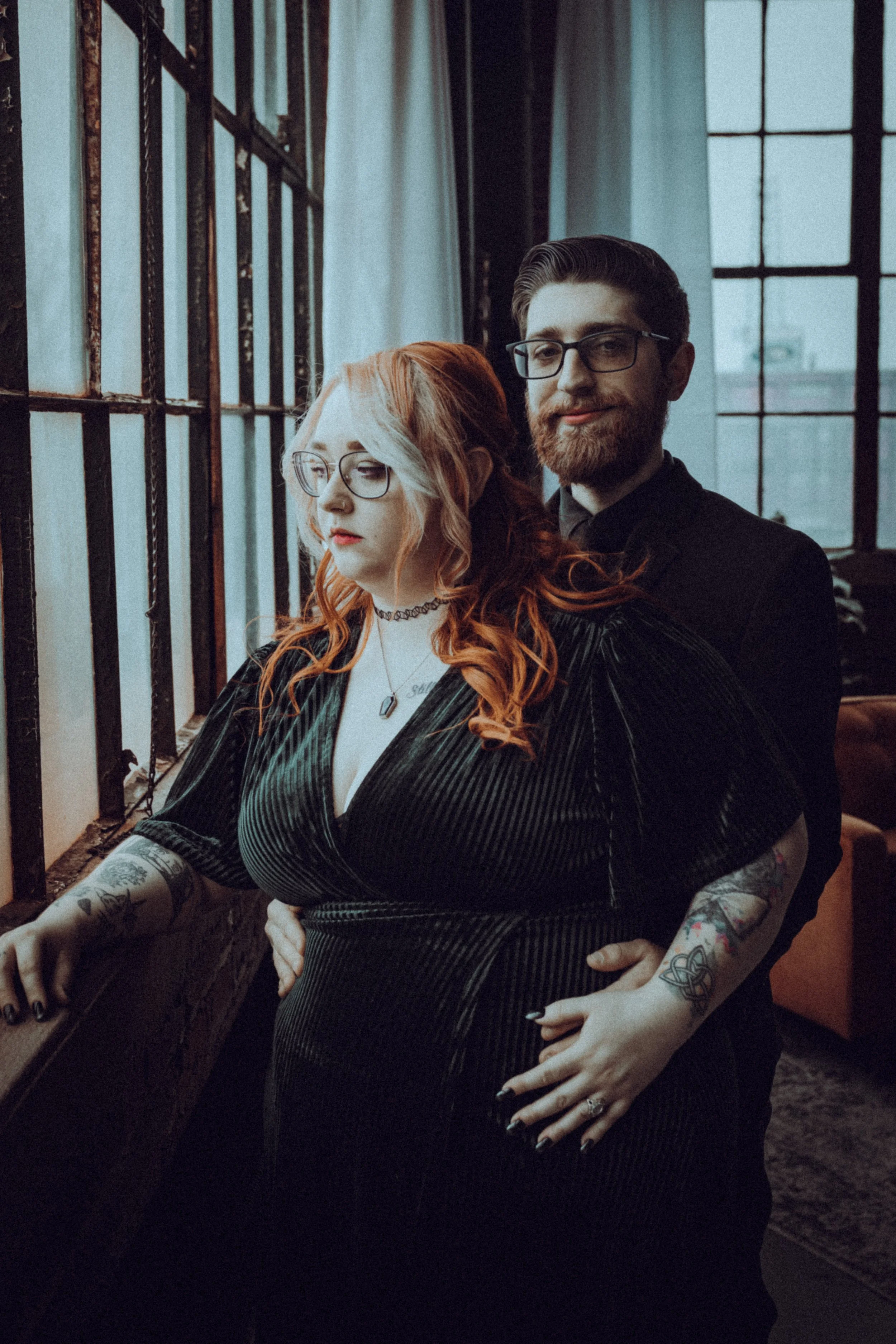 A shirtless man with glasses and a beard stands behind a pregnant woman with red hair, glasses, and tattoos on her arms. They are indoors near a large window with metal bars and light curtains.