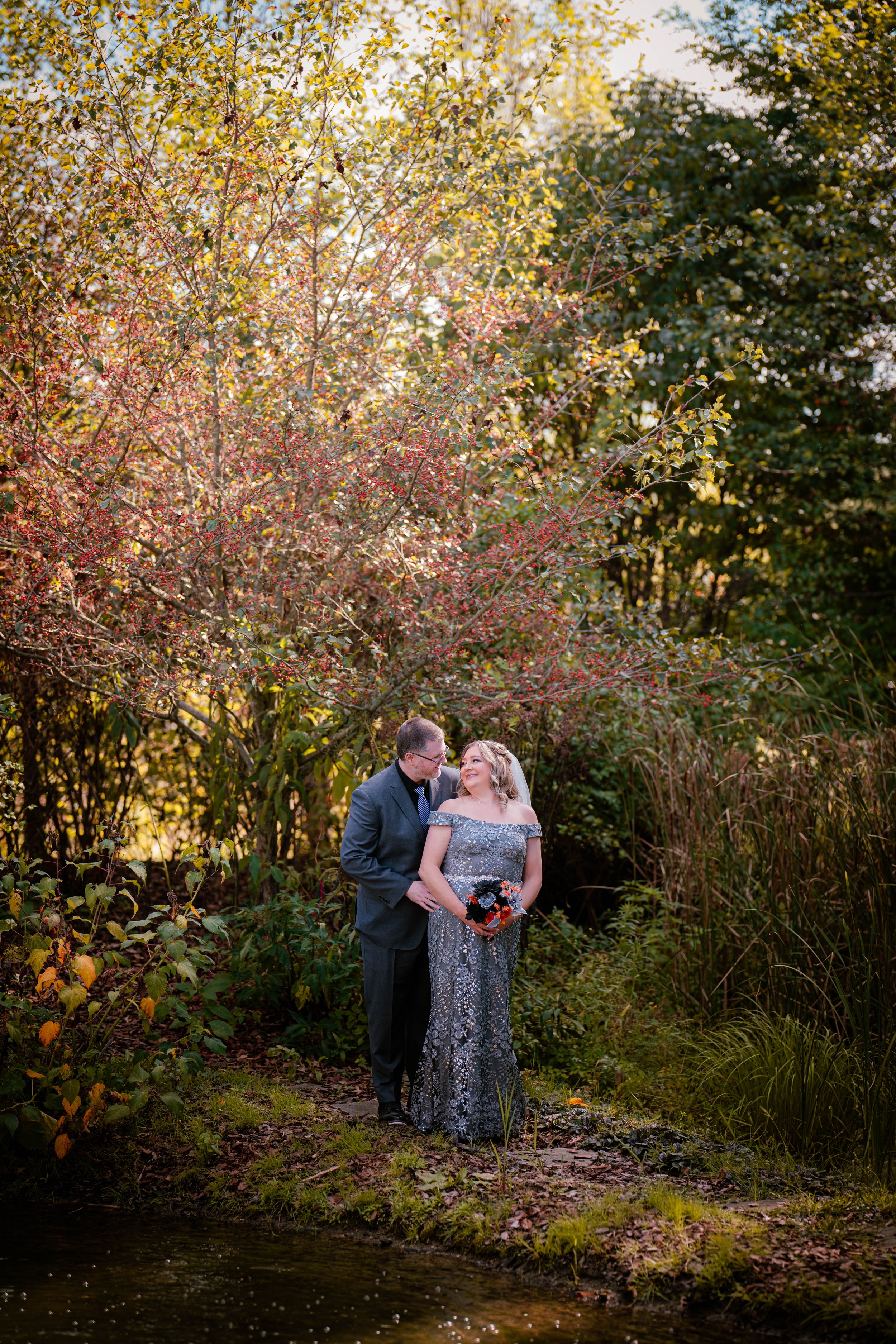 A couple standing near a body of water, surrounded by fall foliage, with the woman holding a bouquet of flowers and the man in a suit, sharing a moment.
