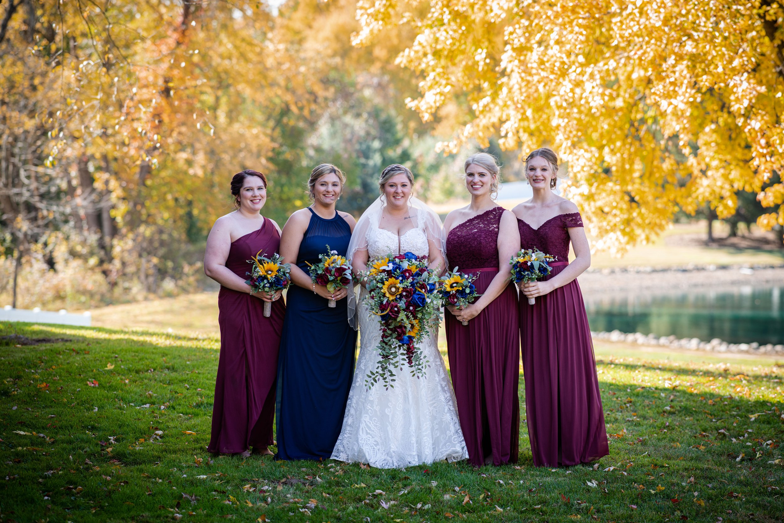A bride in a white wedding gown standing with four bridesmaids in colorful dresses holding bouquets, outdoors in a park with autumn foliage and a pond in the background.