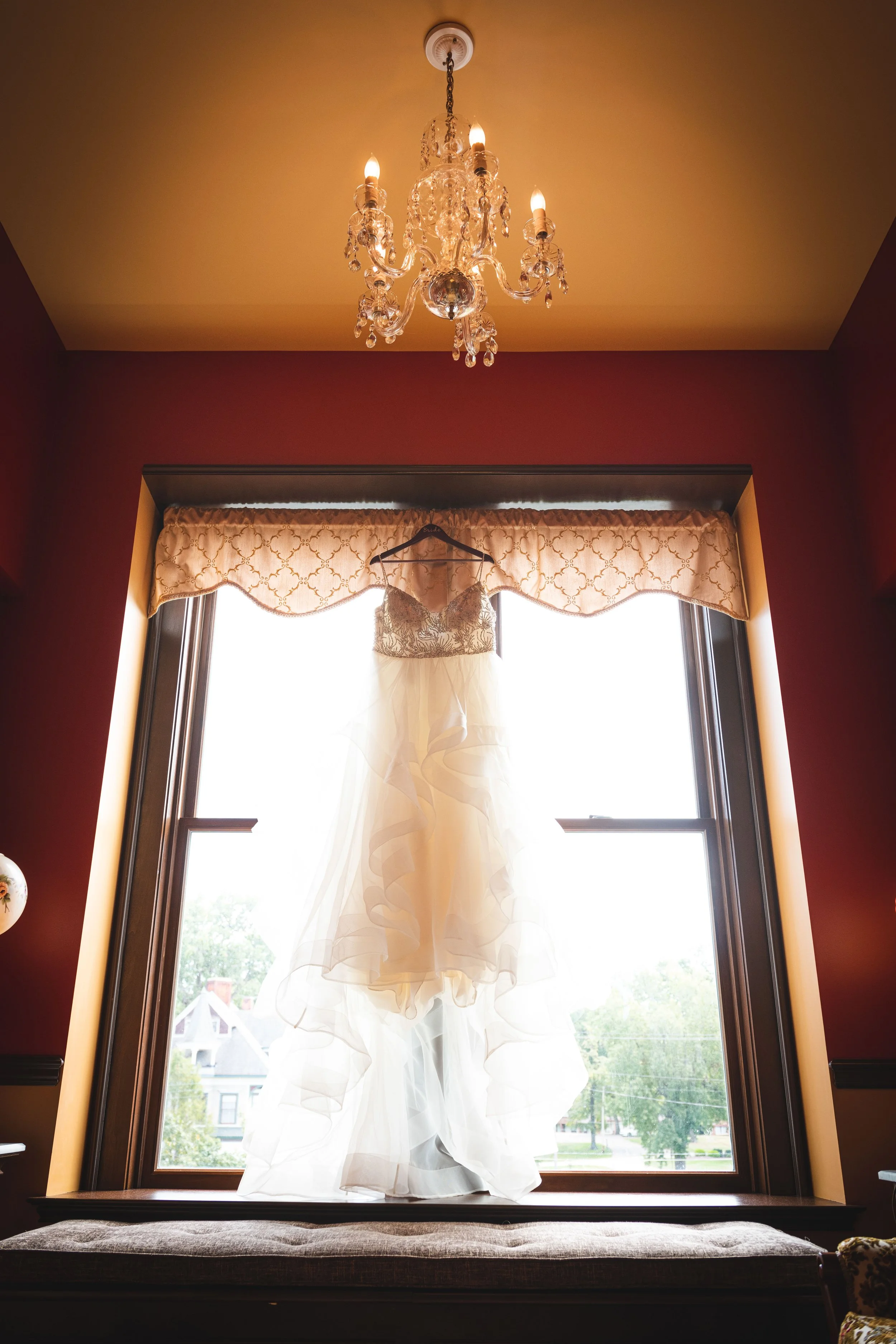 A wedding dress hanging in front of a bright window with a chandelier above.
