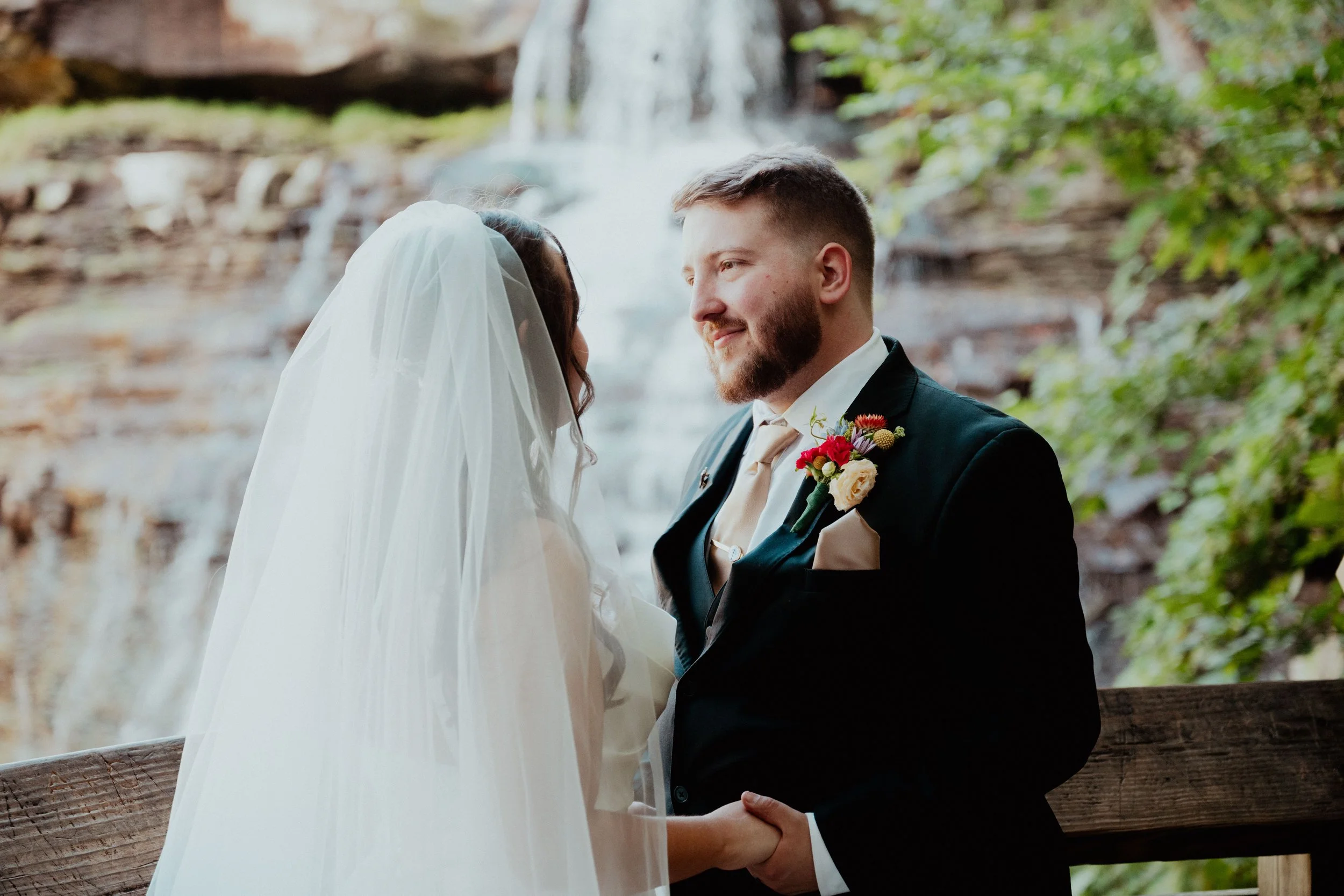 A bride and groom holding hands and gazing at each other outdoors with a waterfall and trees in the background.