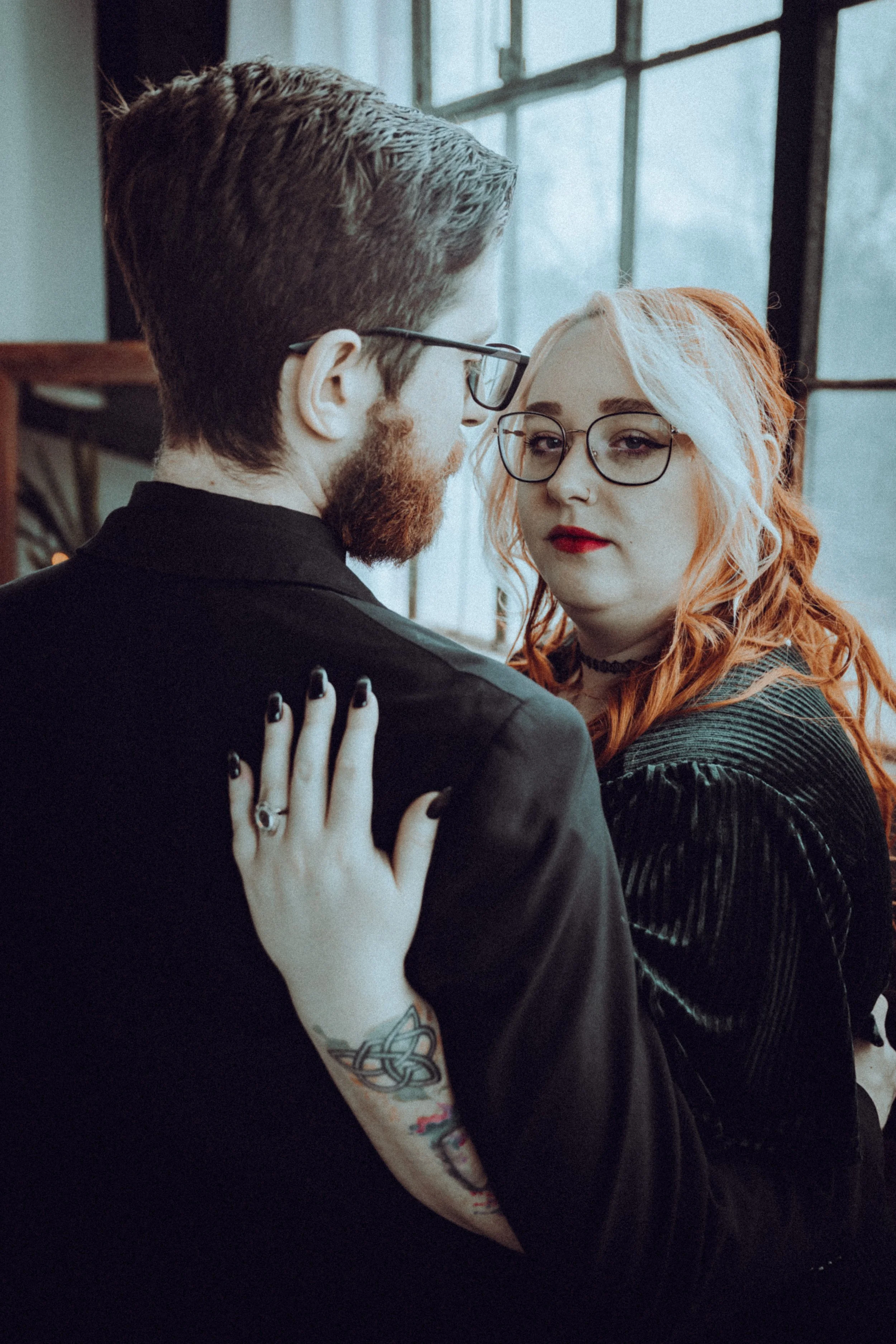 A young man with glasses and a beard and a young woman with glasses and dyed blonde hair are close together, touching foreheads, in an intimate pose, inside near a window.