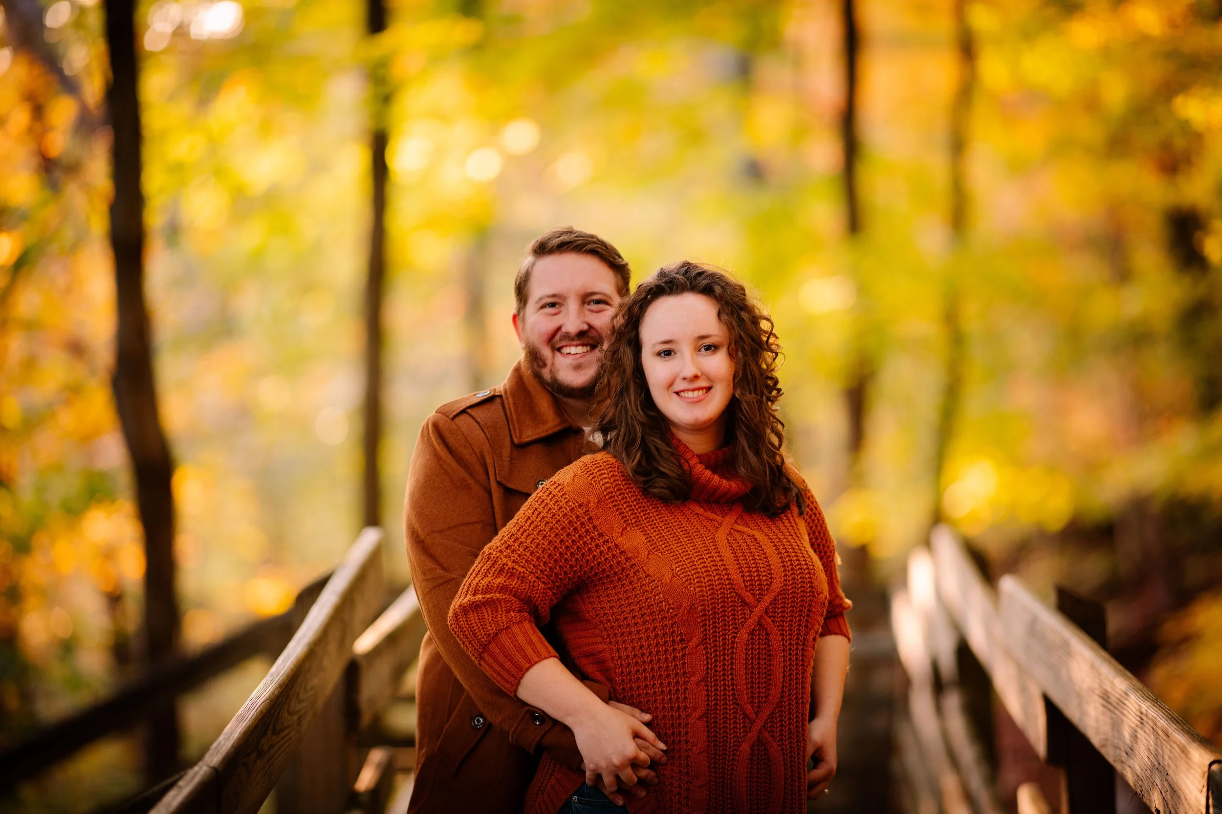 A couple standing on a wooden bridge surrounded by autumn trees with yellow and orange leaves.