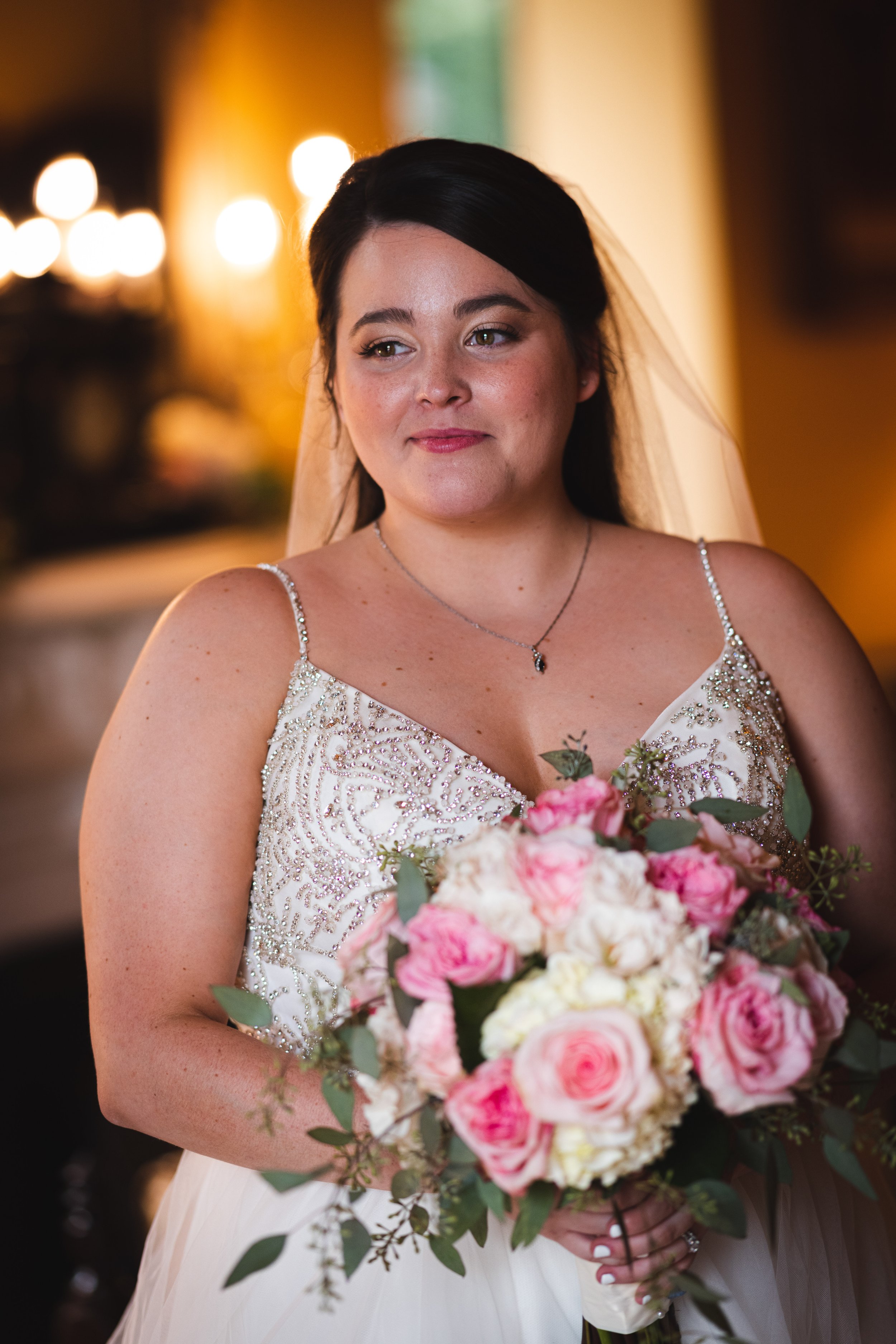 A bride holding a bouquet of pink and white roses, wearing a wedding dress with beaded details and a veil, standing indoors with warm lighting.