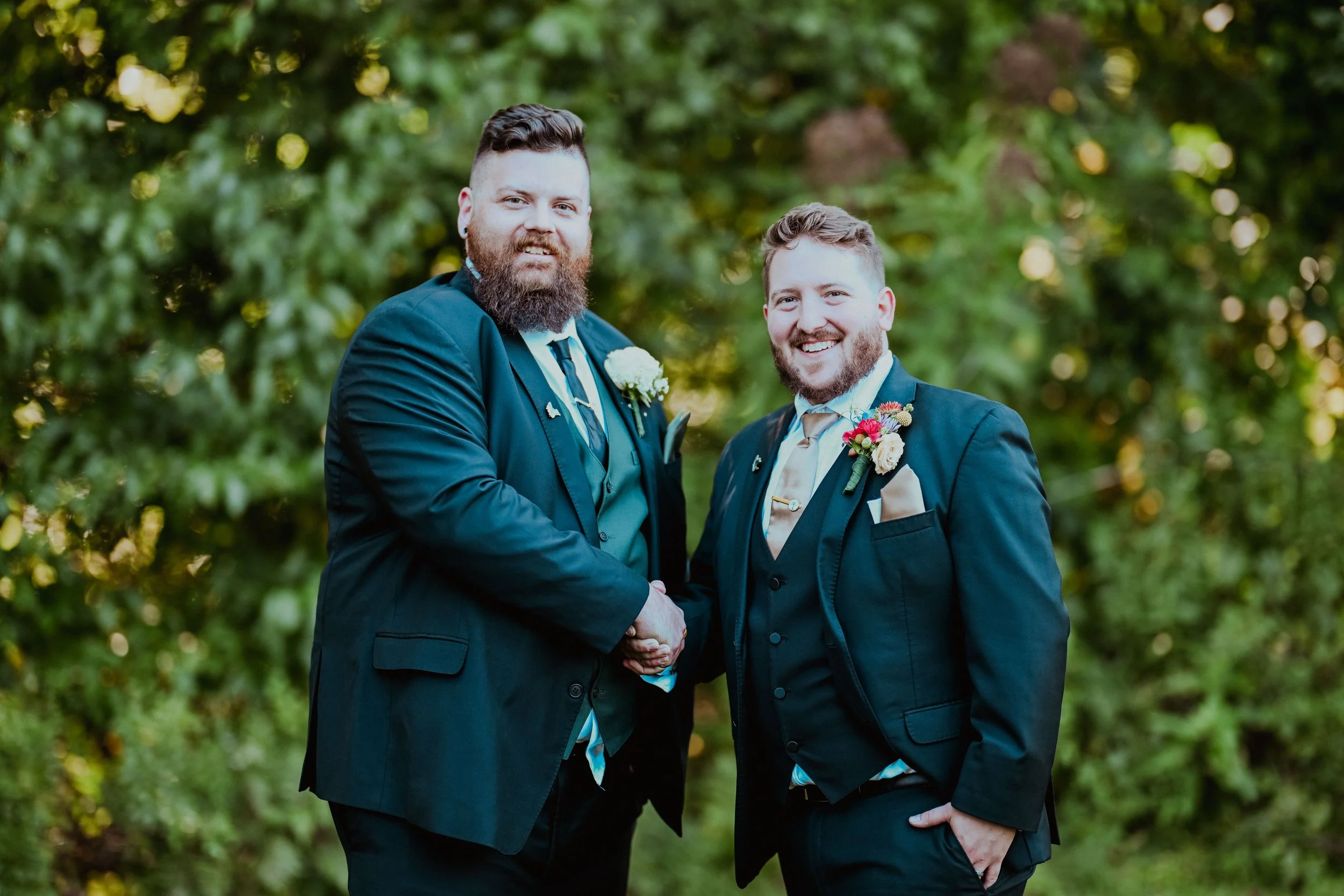 Two men in formal suits with boutonnières shaking hands outdoors, smiling at camera, surrounded by green trees.