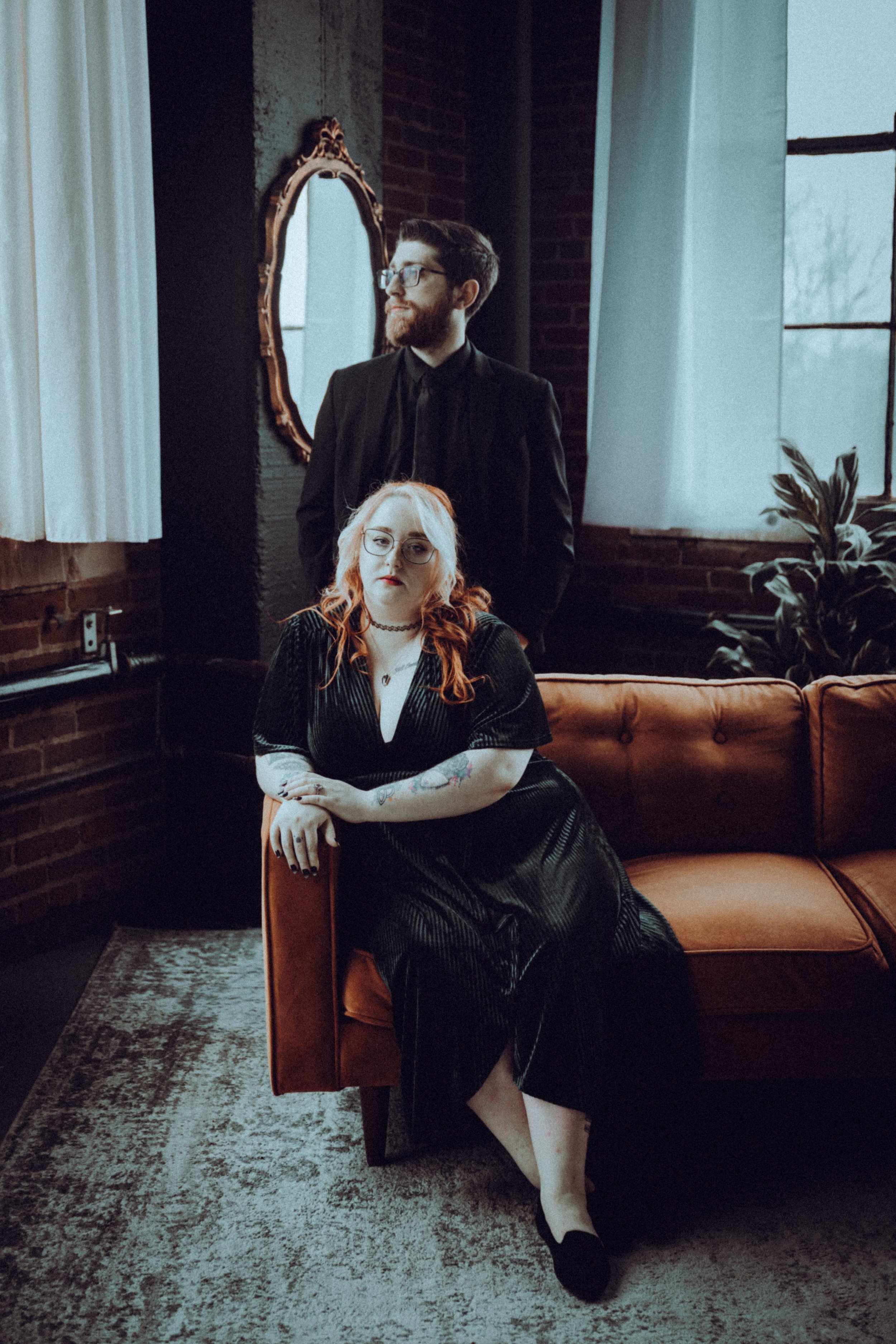 A man standing behind a woman who is sitting on an orange couch in a dimly lit room with brick walls, large windows, and a plant.