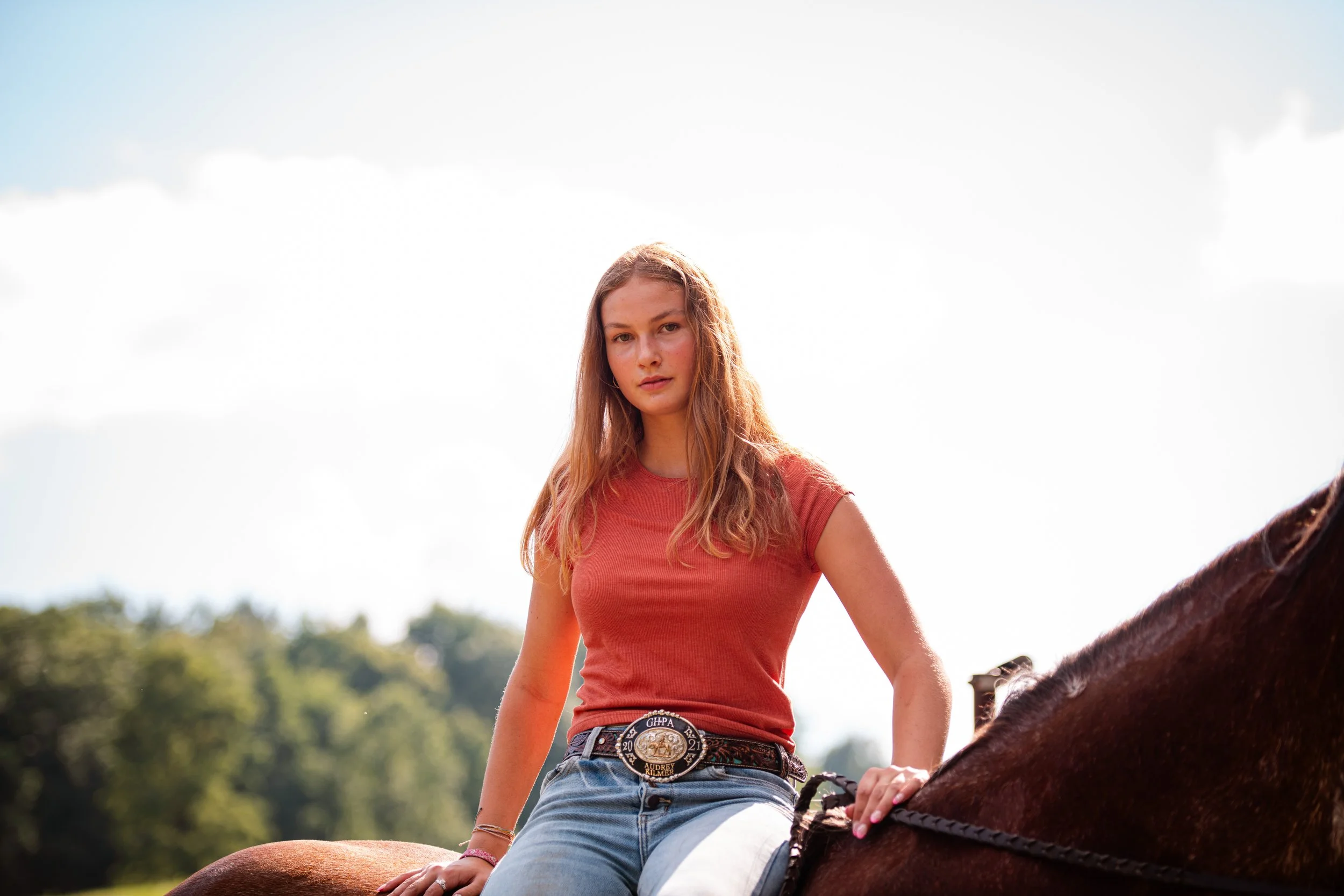 Young woman with wavy hair wearing a red t-shirt and jeans riding a brown horse outdoors on a sunny day.