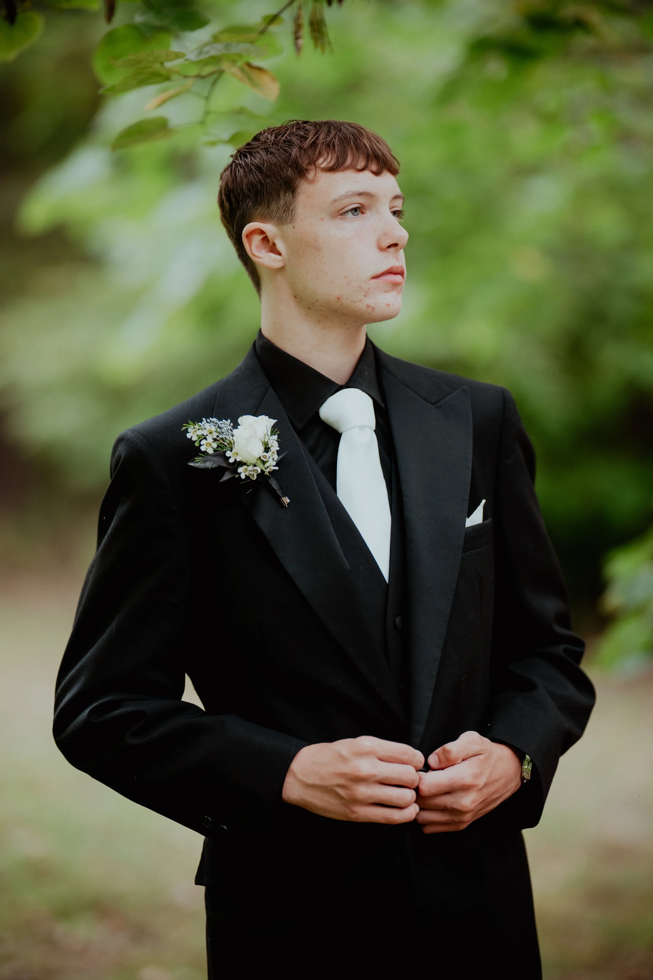 Young man wearing a black tuxedo with a white tie and boutonniere, standing outdoors surrounded by greenery.