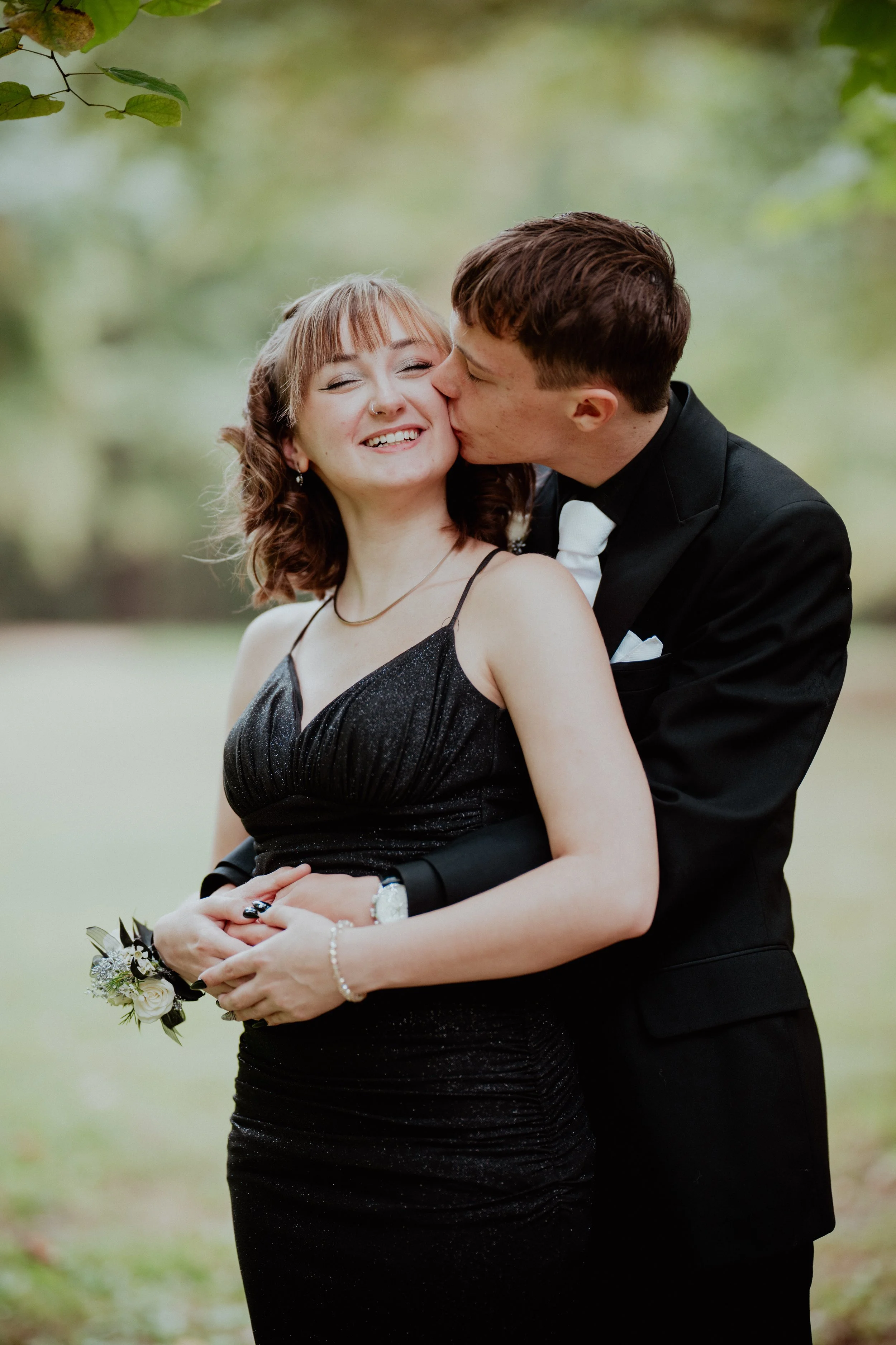 A young woman in a black, sparkly dress is smiling as a young man in a black tuxedo kisses her cheek in an outdoor setting with green blurred background.