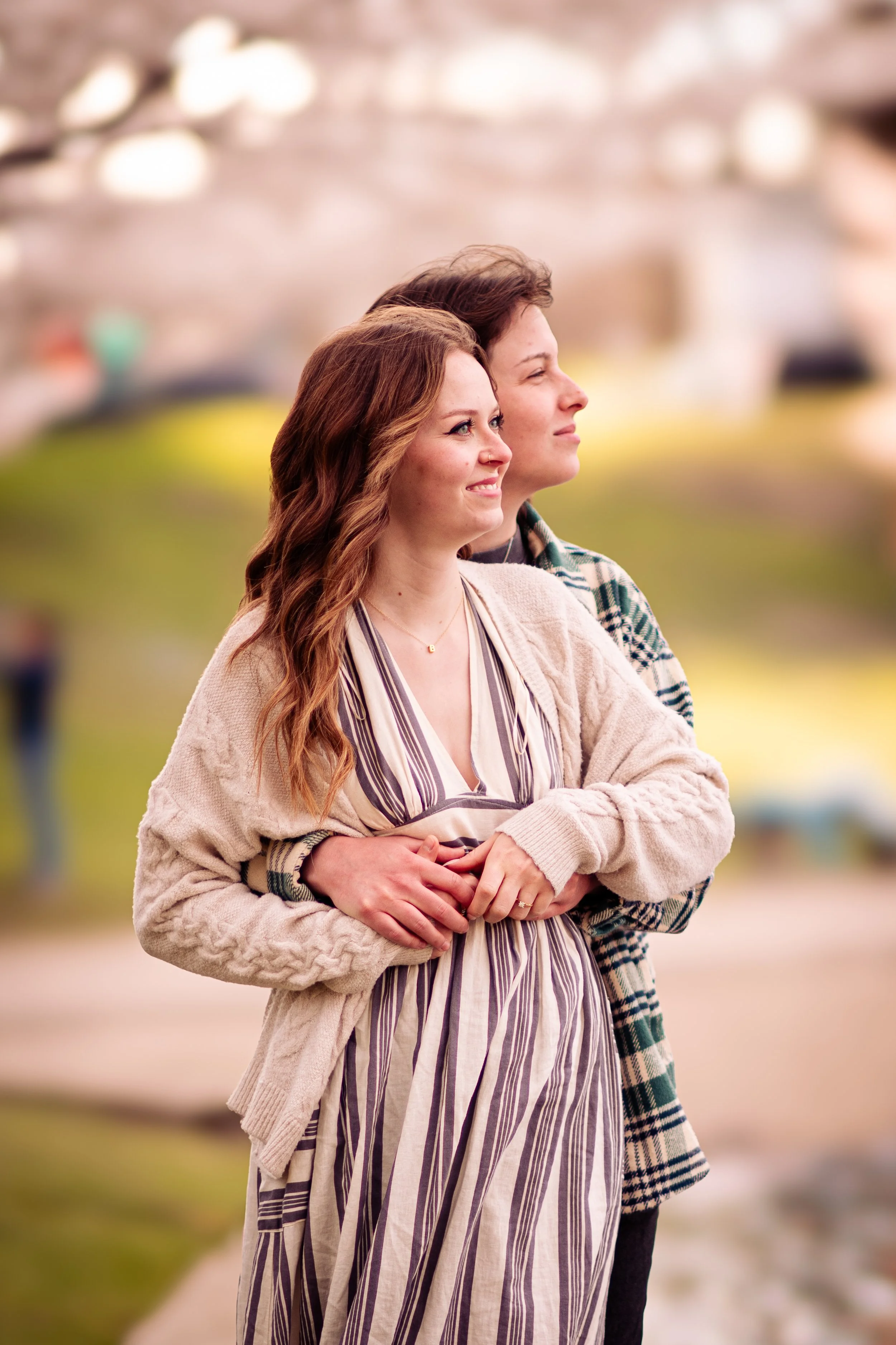 A couple stands outdoors, embracing and looking into the distance. The woman has red hair, and the man has brown hair. The background is blurred with hints of trees and a park setting.