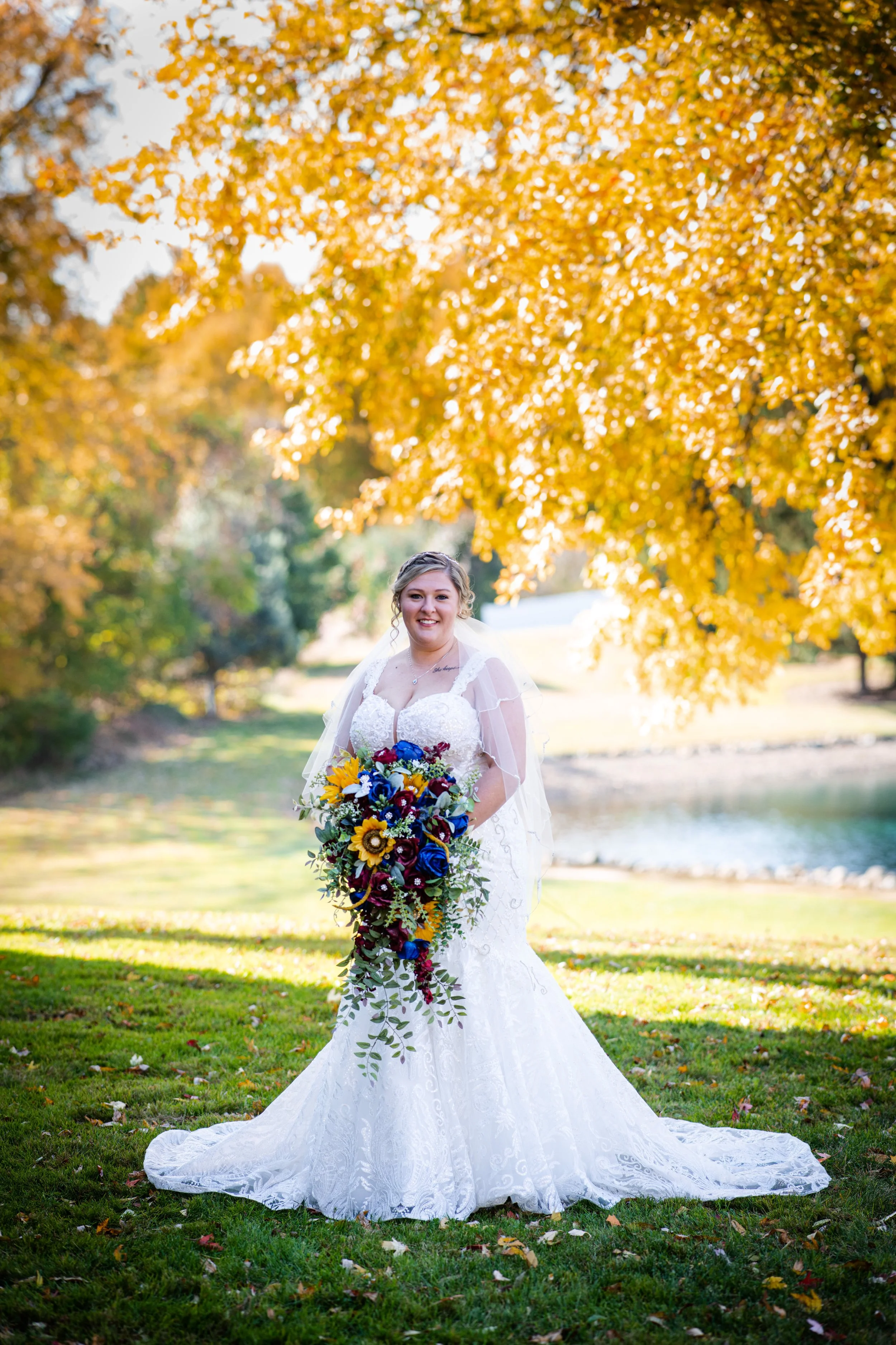 Bride in a white wedding dress holding a colorful bouquet outdoors during autumn with yellow-orange trees and a pond in the background.