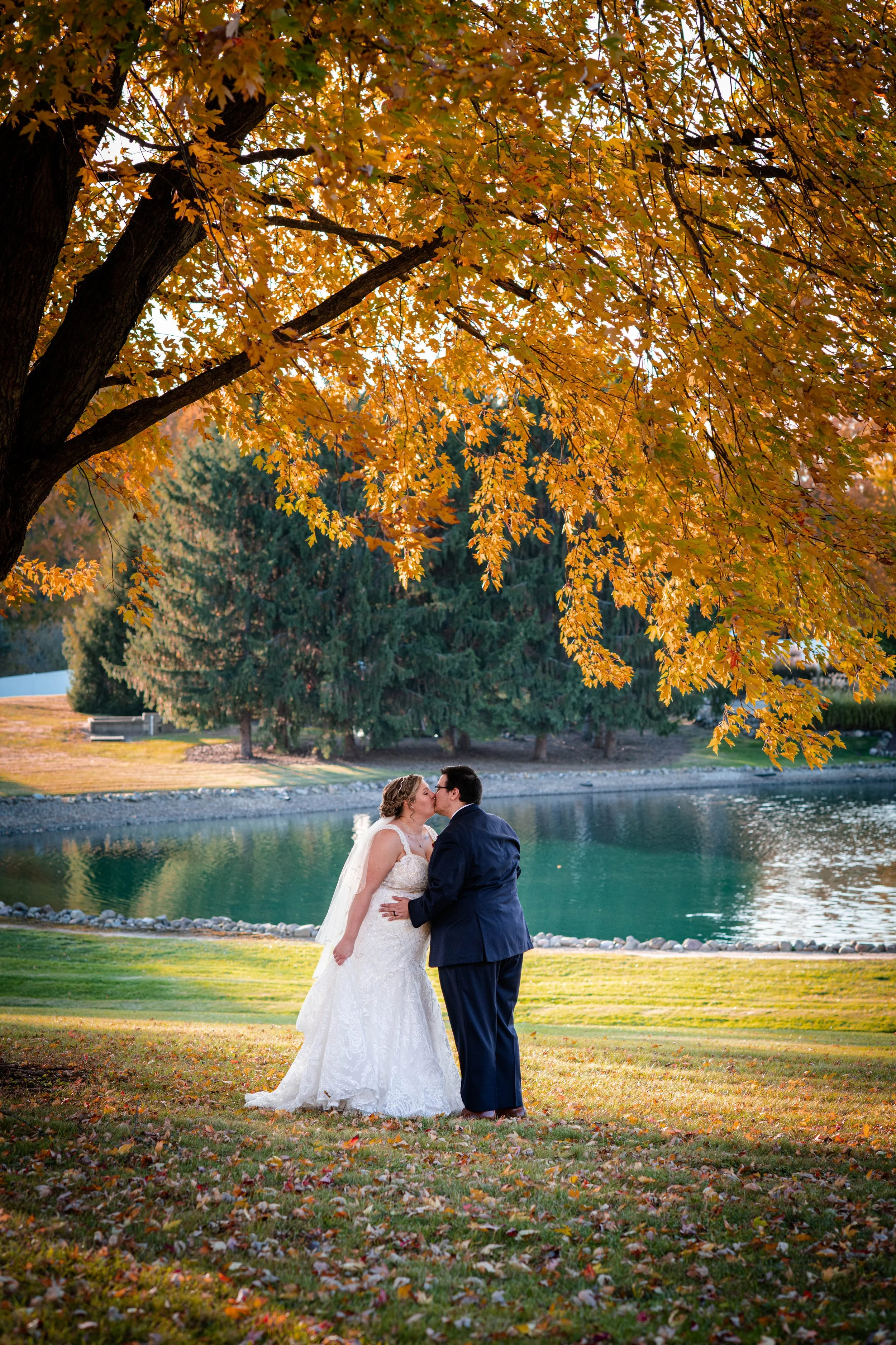 A bride and groom kissing outdoors by a lake under a large tree with orange autumn leaves.