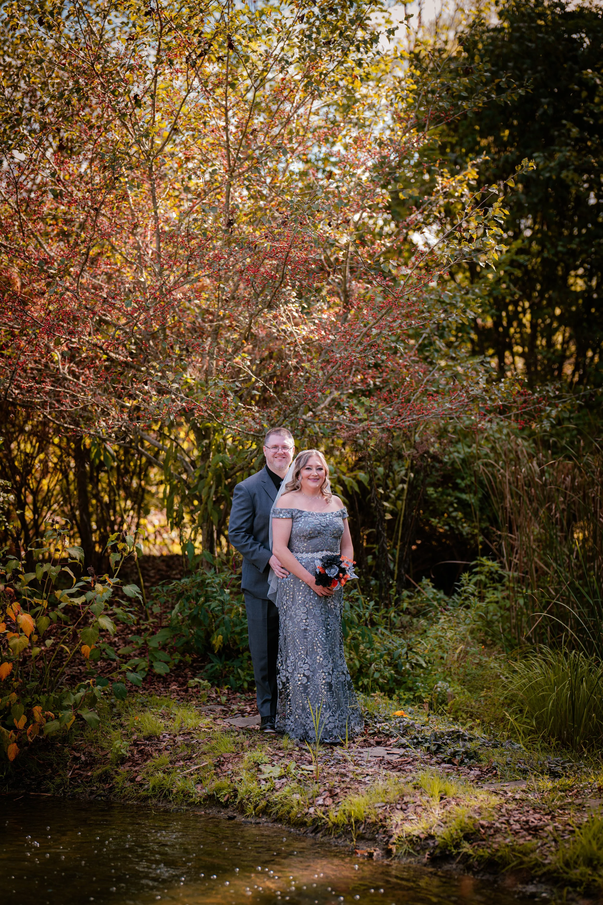 A couple dressed in formal attire standing on a grassy area near a small body of water, surrounded by trees with autumn foliage.