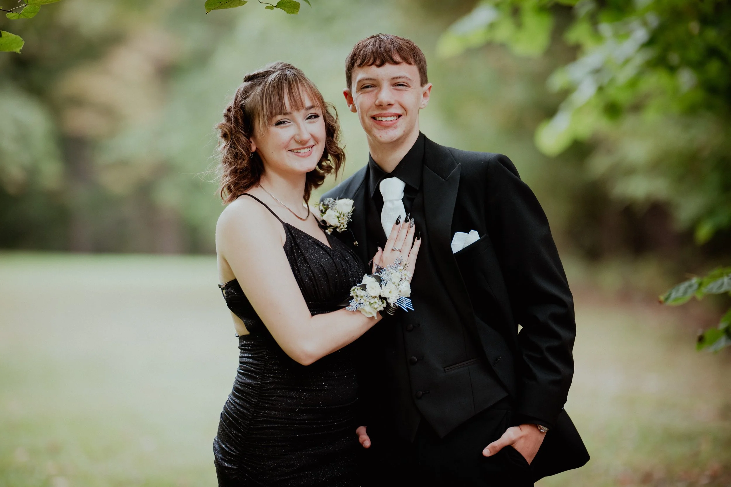A young couple dressed in formal attire, smiling outdoors during daytime with greenery in the background. The woman is wearing a black dress with spaghetti straps and has flowers on her wrist, while the man is in a black suit with a white tie and flo