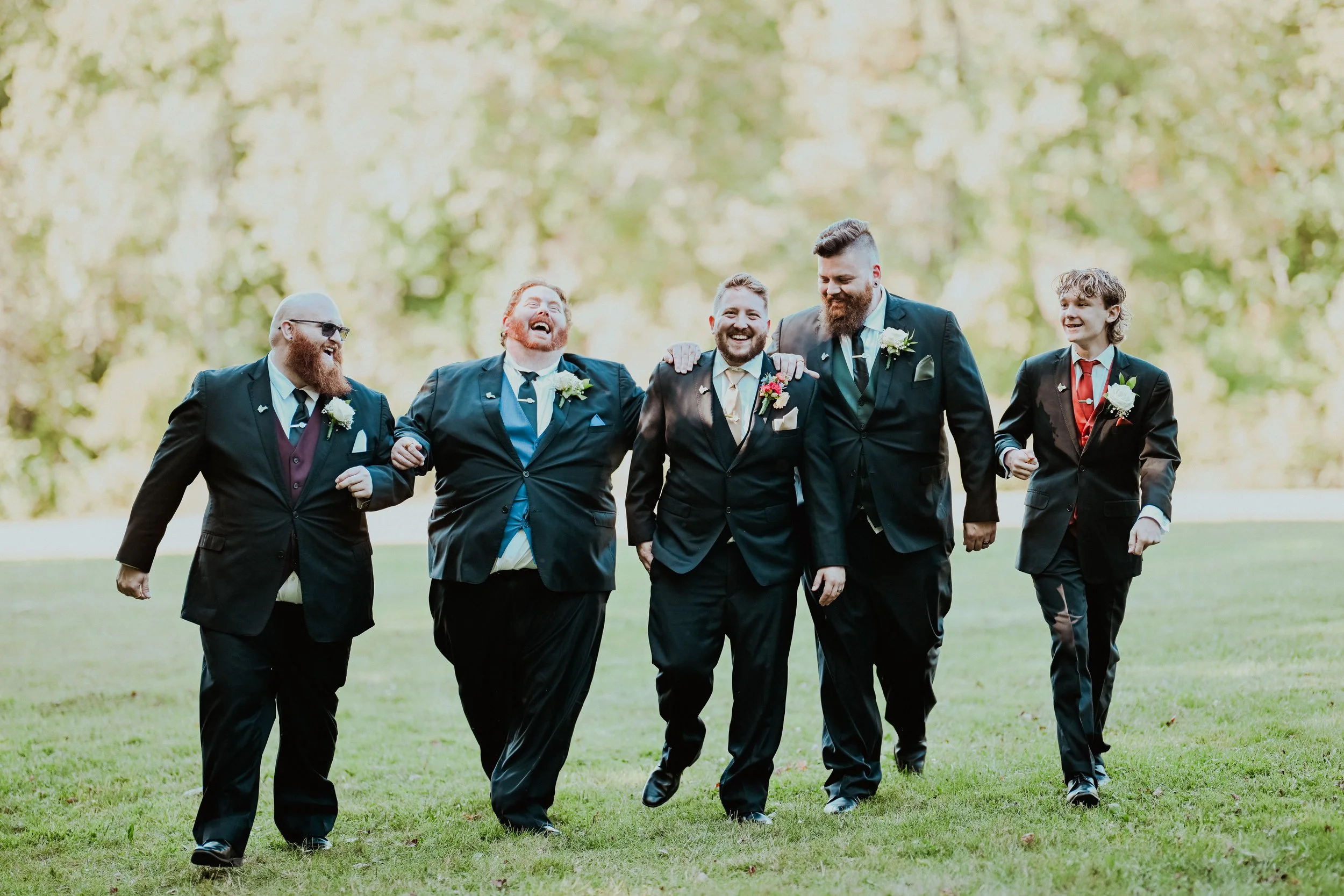 A group of men in suits walking together on a grassy field during a wedding, smiling and enjoying each other's company, with green trees in the background.