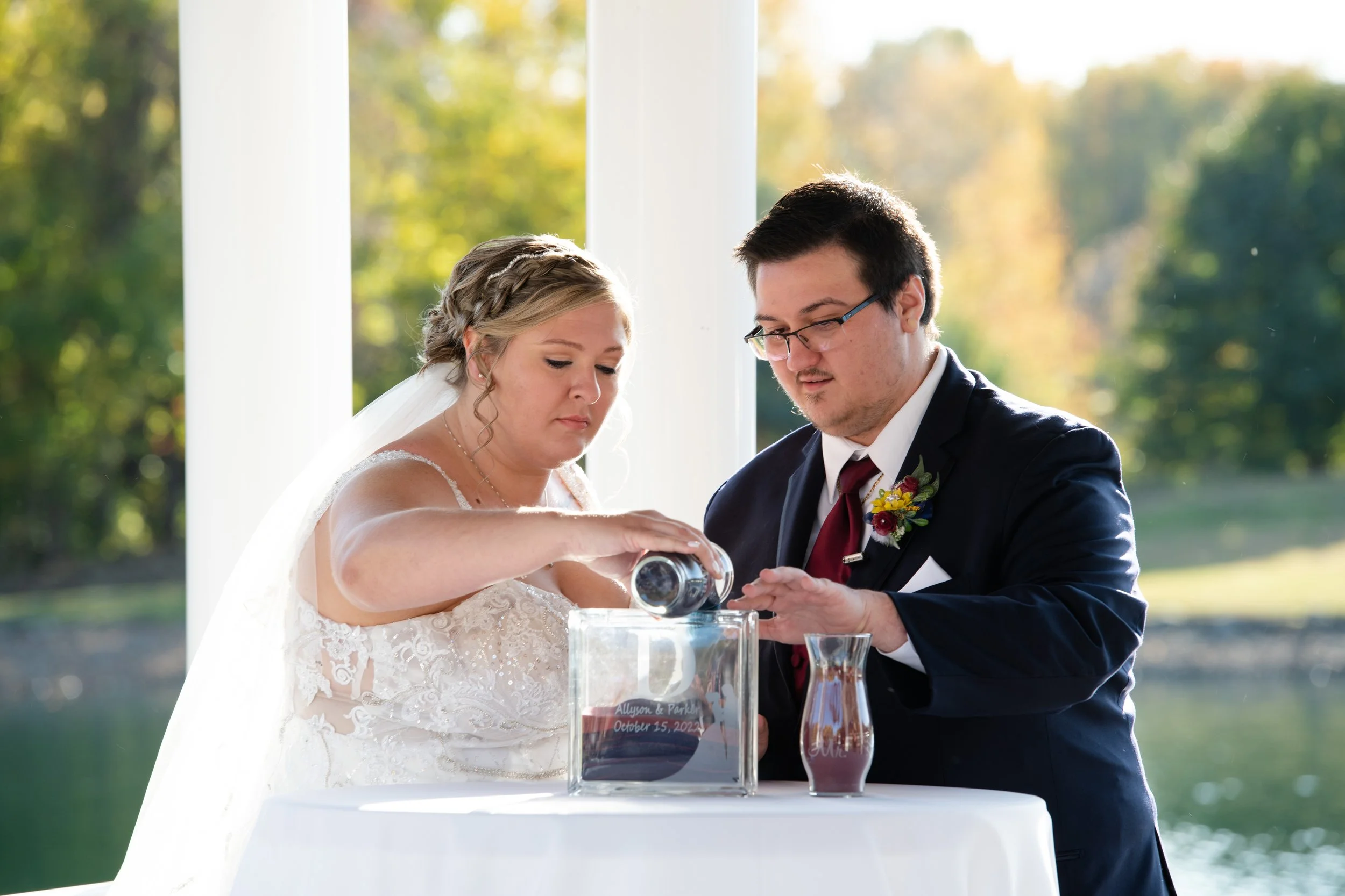 Bride and groom participating in a sand ceremony during their wedding, pouring sand into a glass vessel outdoors with a lake and trees in the background.