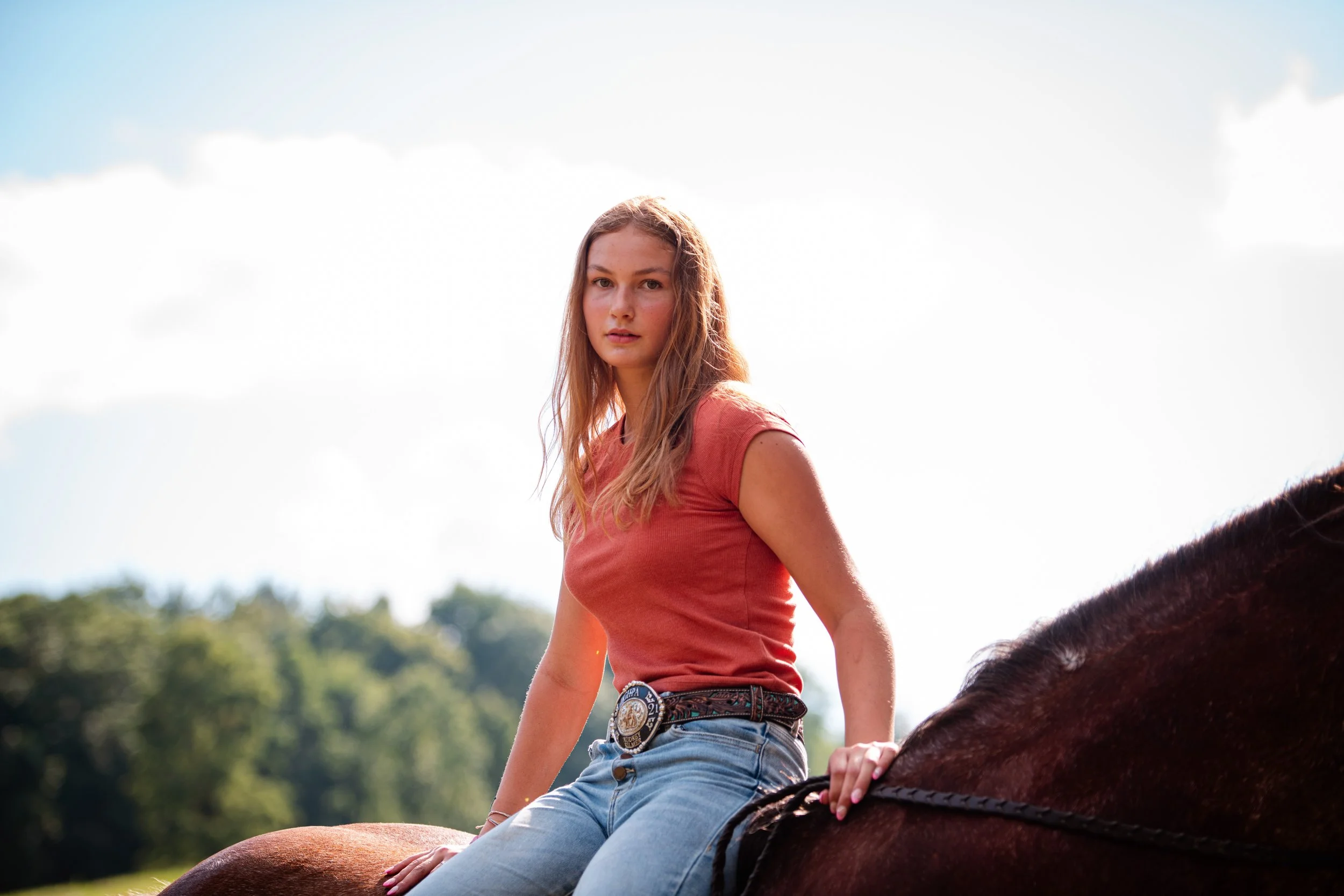 Young woman riding a horse outdoors on a sunny day with trees in the background.