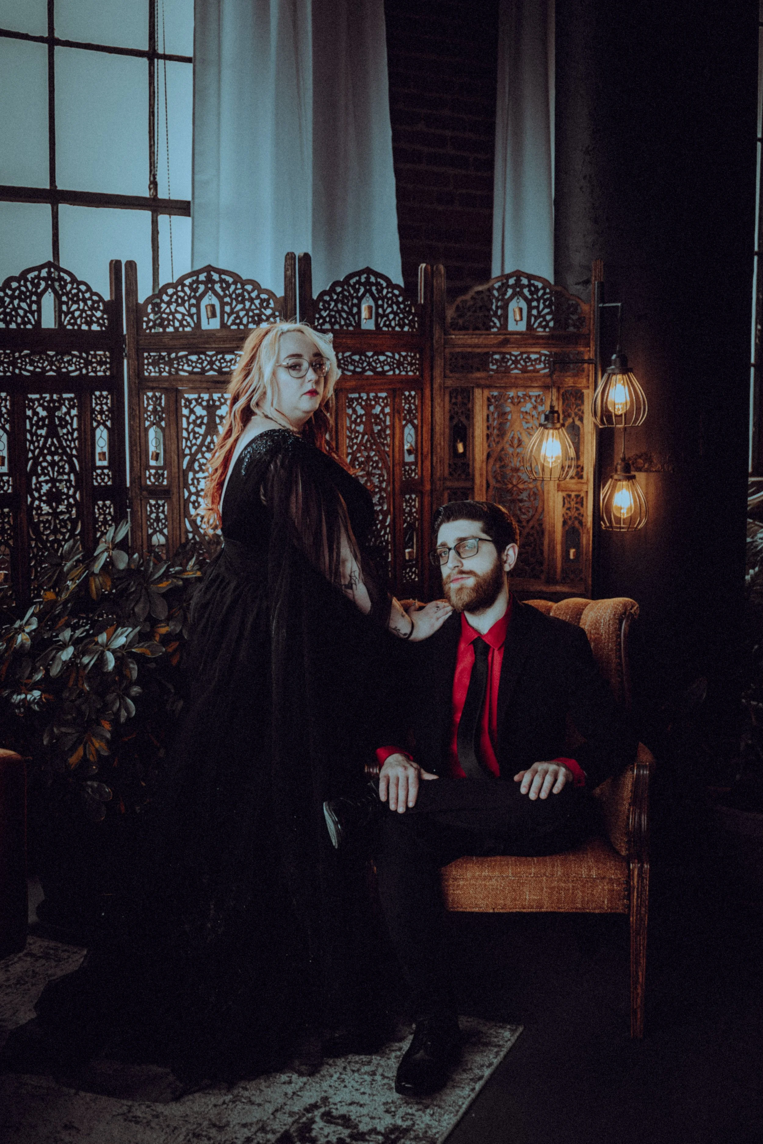 A woman with long, wavy blonde hair and glasses, dressed in a black gown, standing next to a seated man with dark hair, a beard, glasses, wearing a black suit with a red shirt and tie. They are in a dimly lit room with a decorative wooden room divide