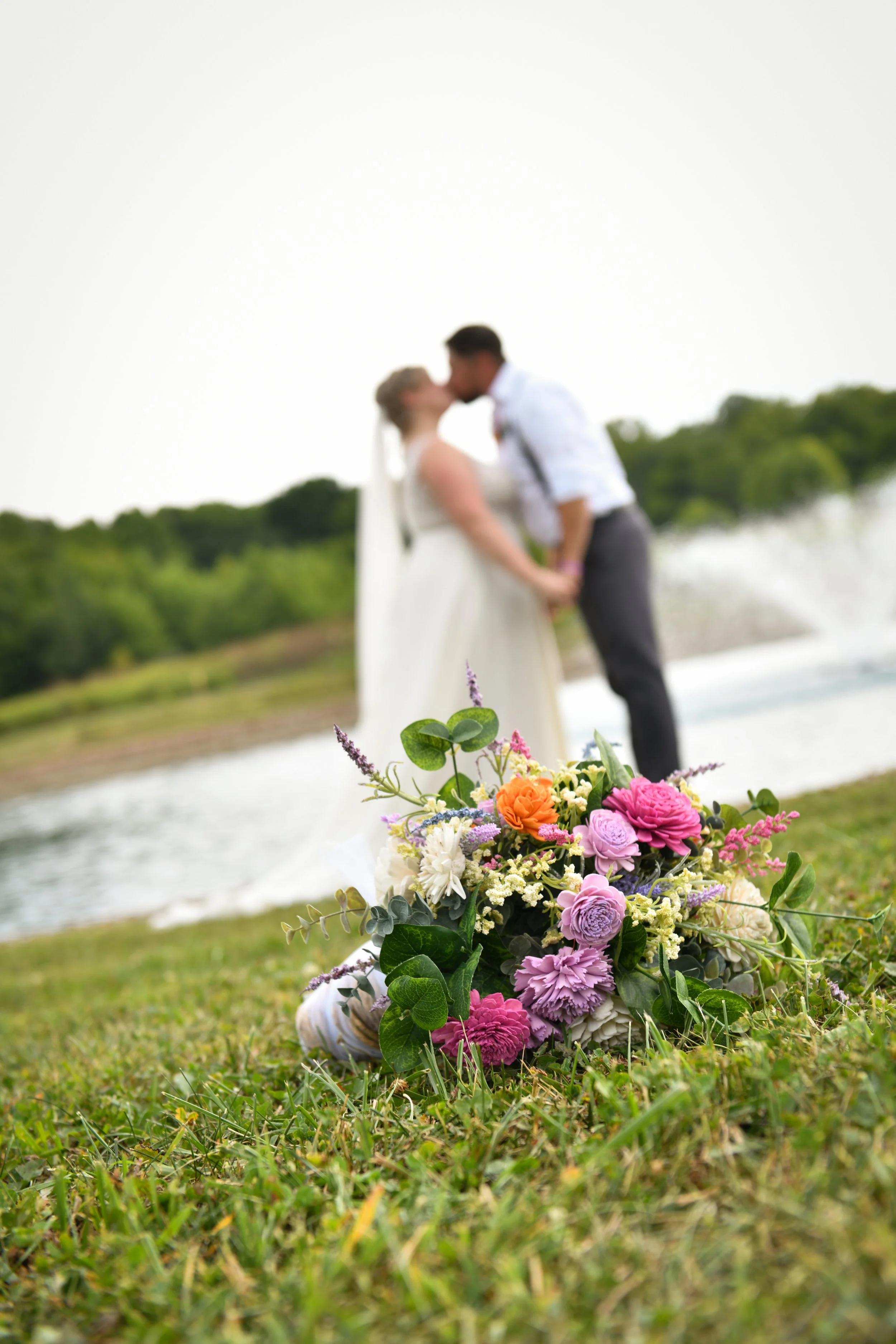Colorful wedding bouquet in focus with a bride and groom kissing in the background near a lake or pond