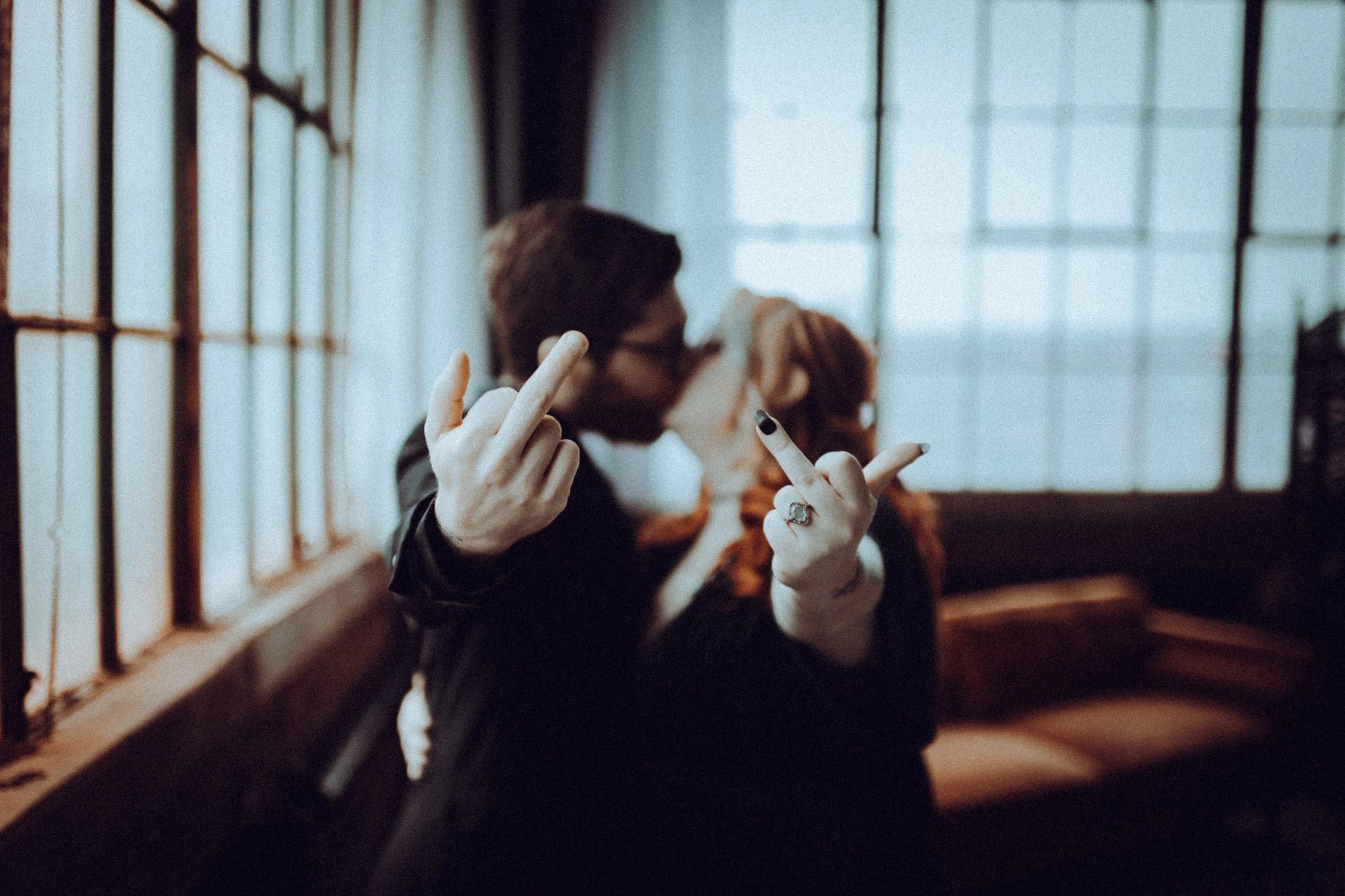 A person with dark hair and glasses makes a rude gesture with both hands, middle fingers raised, in an industrial-style room with large windows, while kissing a person with red hair in the background.