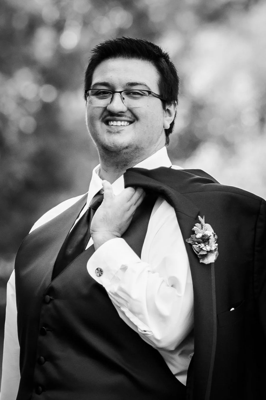 A young man with glasses in a tuxedo, smiling, holding a suit jacket over his shoulder, outdoors in a natural setting, black and white photograph.