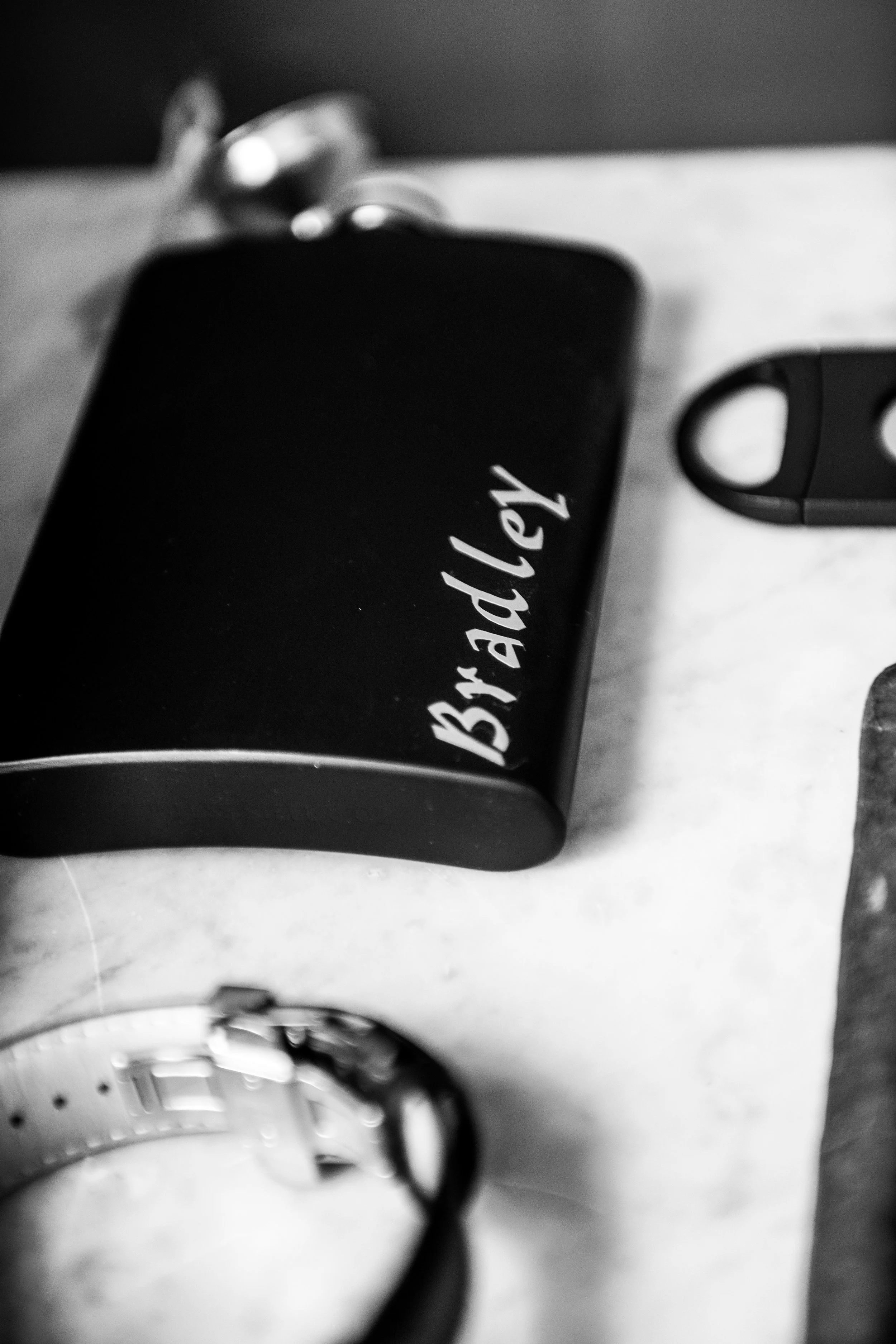 Close-up of a black bottle with the label "Bradley" on a white surface, with a watch, a key fob, and a pair of earbuds partially visible around it.