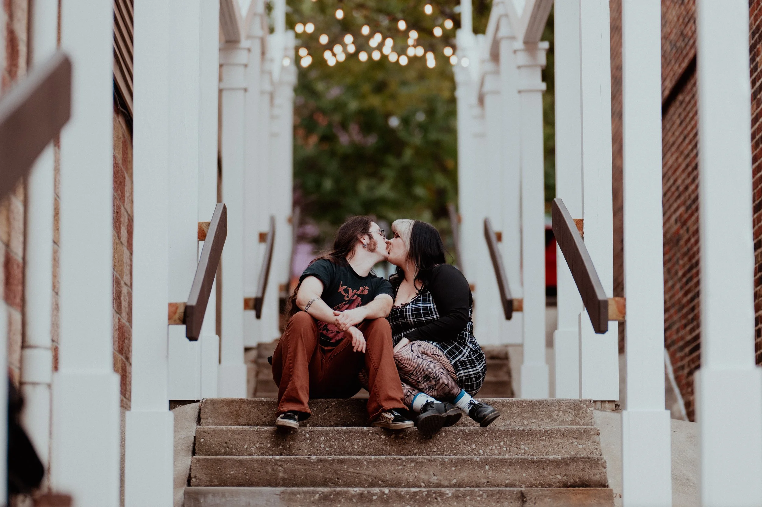Two women sitting on outdoor steps sharing a kiss with string lights overhead and trees in the background.