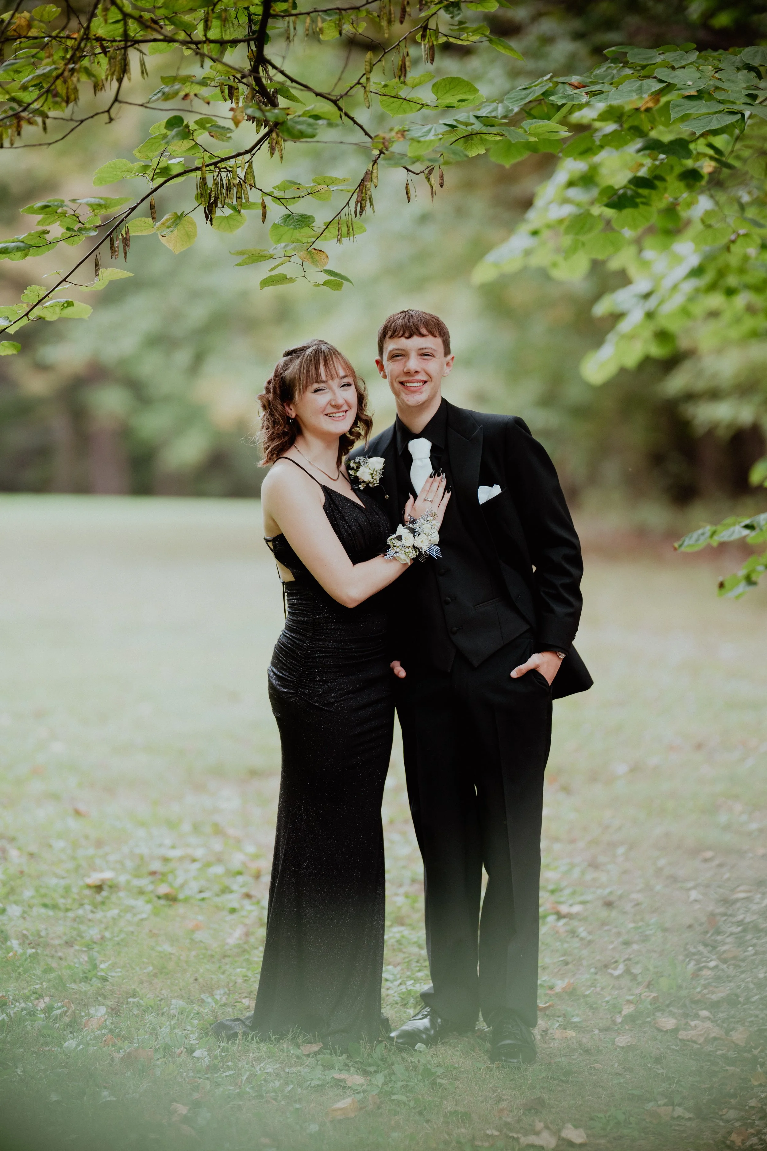 A smiling young couple dressed in formal attire standing together outdoors in a park-like setting with trees and grass, posing for a photo.