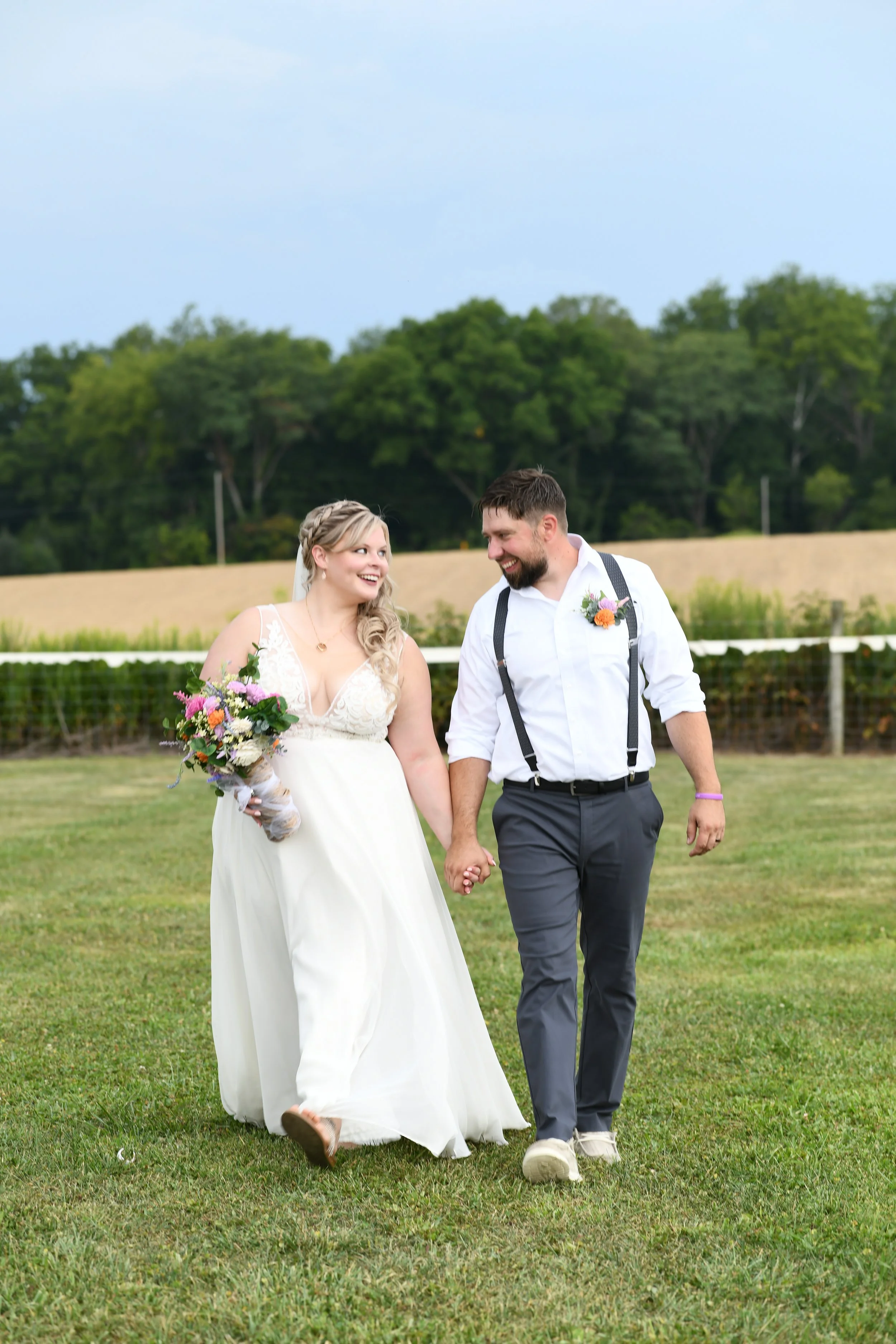 A newly married couple walking hand in hand on a grassy field, smiling at each other during their wedding photo shoot. The bride is holding a colorful bouquet and wearing a white wedding dress, while the groom is dressed casually with a white shirt, 