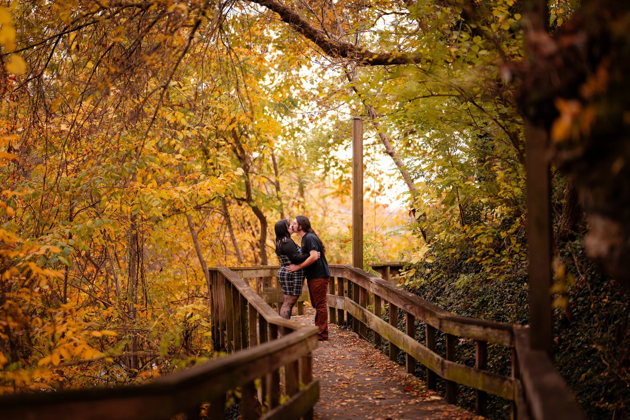 A couple embraces and kisses on a wooden bridge surrounded by colorful autumn trees with yellow and orange leaves.