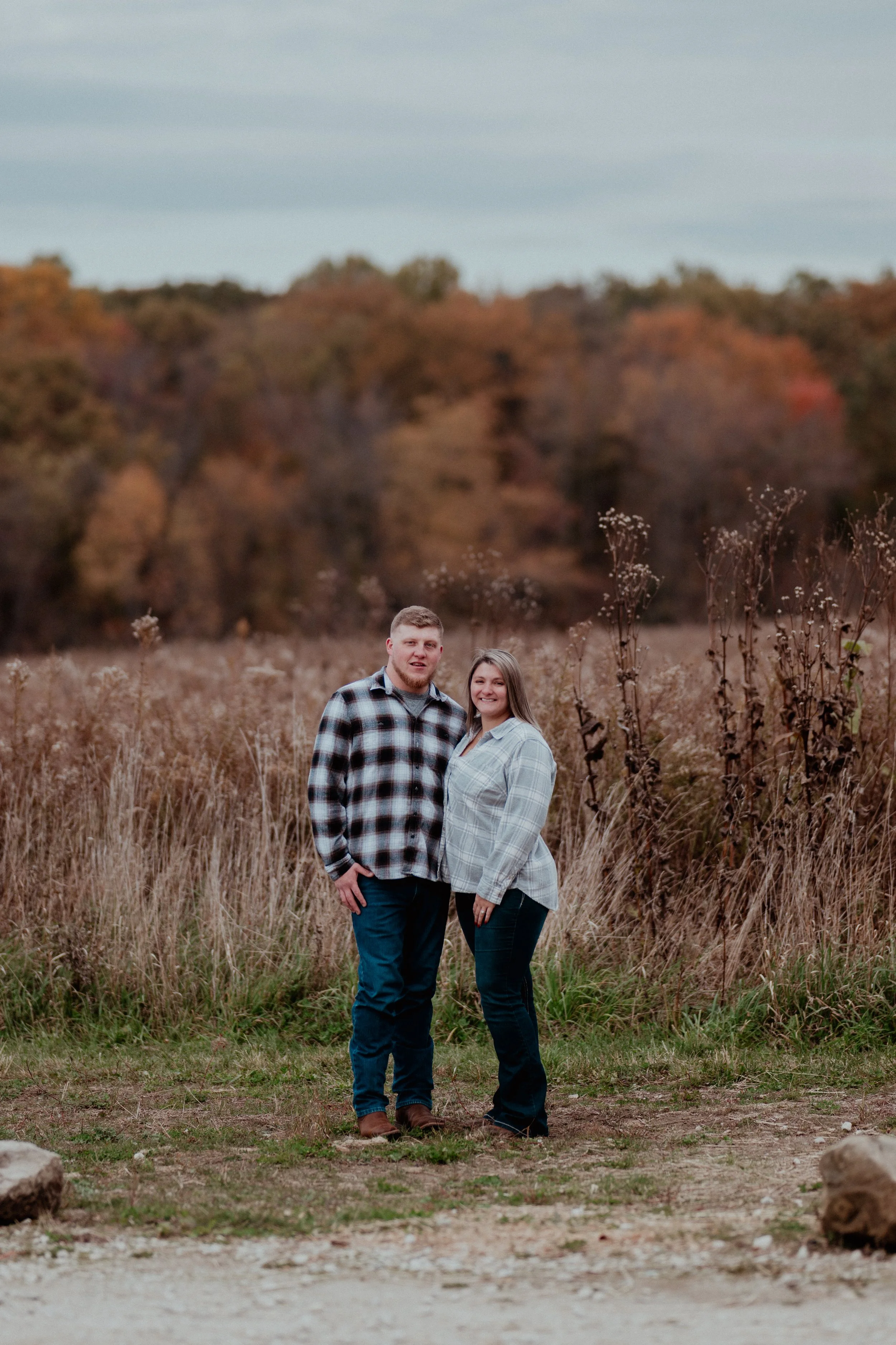 A smiling couple standing together outdoors with autumn foliage in the background.