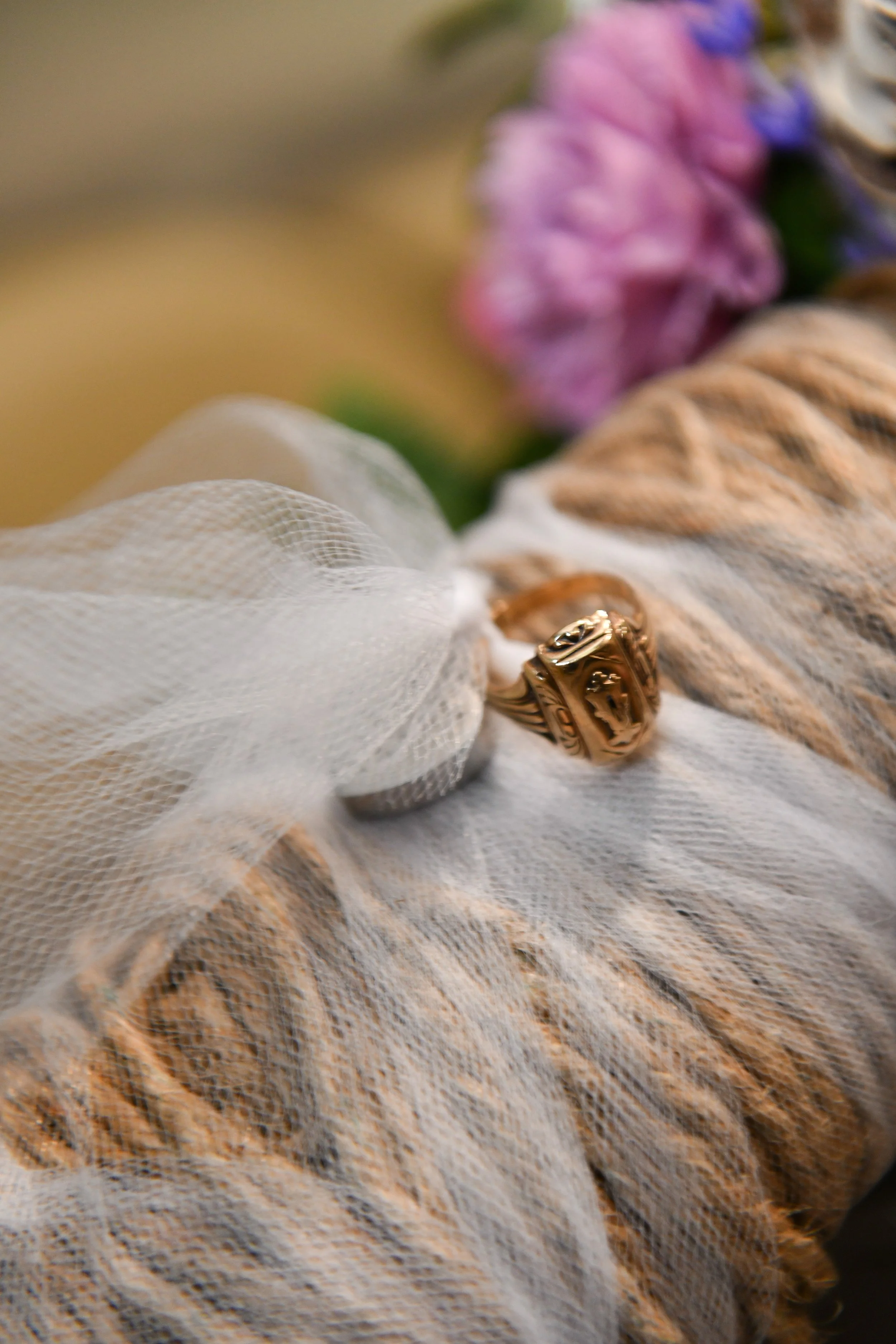 Close-up of a gold ring resting on a piece of gauze fabric, with blurred colorful flowers in the background.