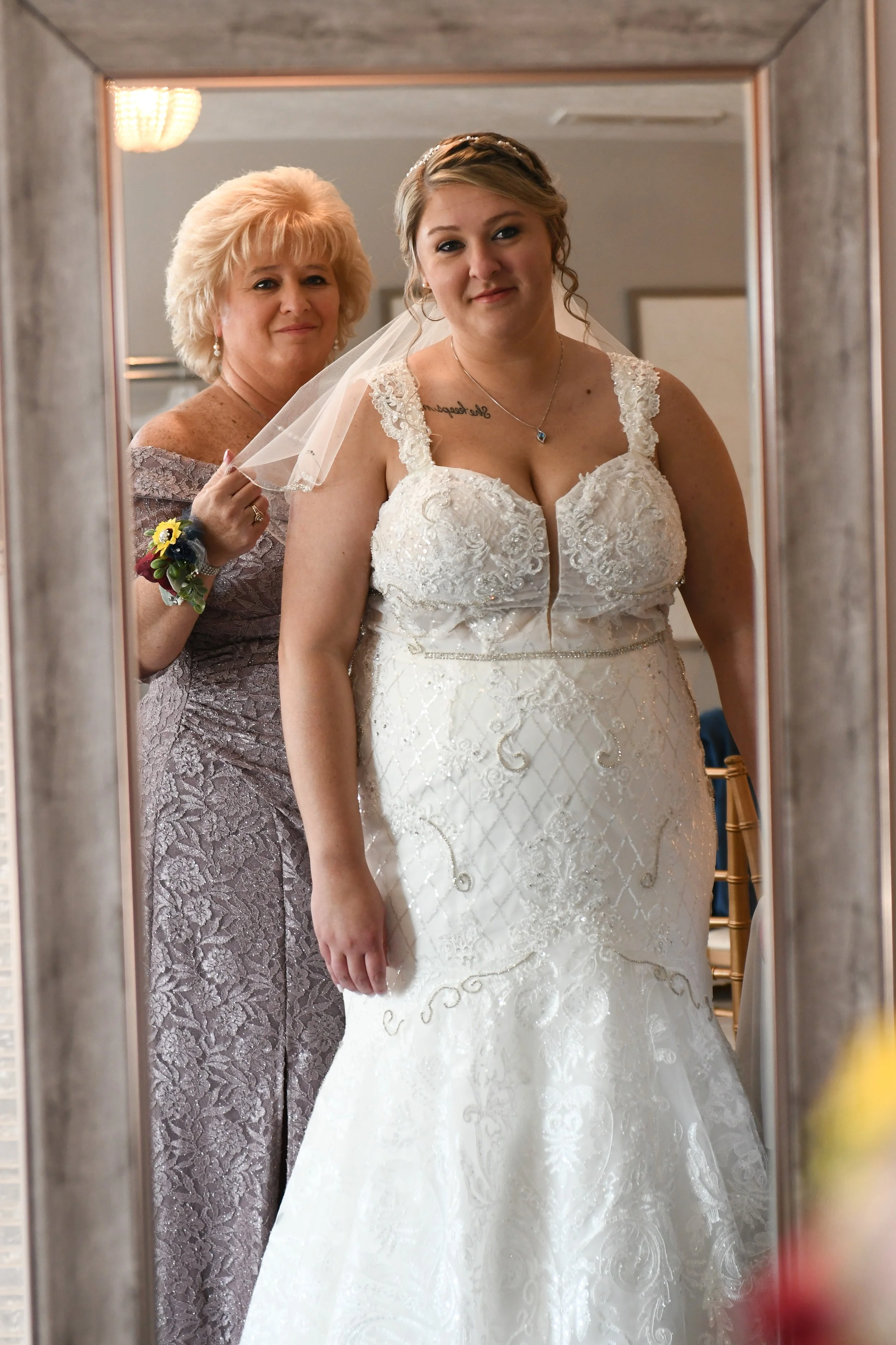 A bride in a white lace wedding gown taking a photo in a mirror, with a woman in a purple dress standing behind her.