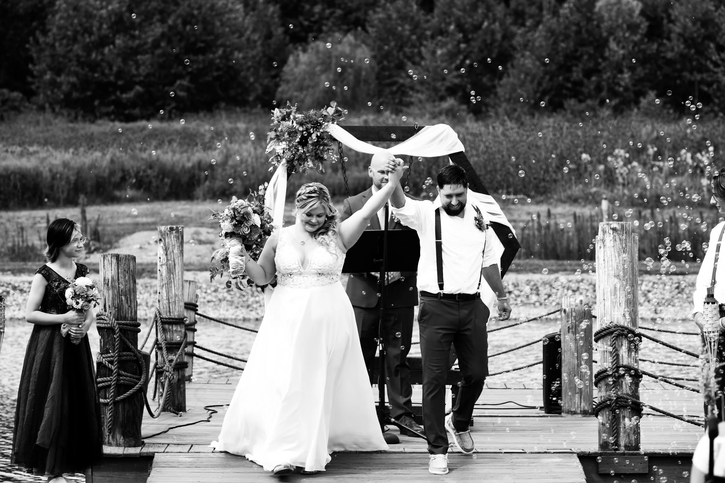 A bride and groom celebrating after their wedding on a wooden dock near the water, with a flower girl holding flowers on the left and guests on the right, as bubbles float around.
