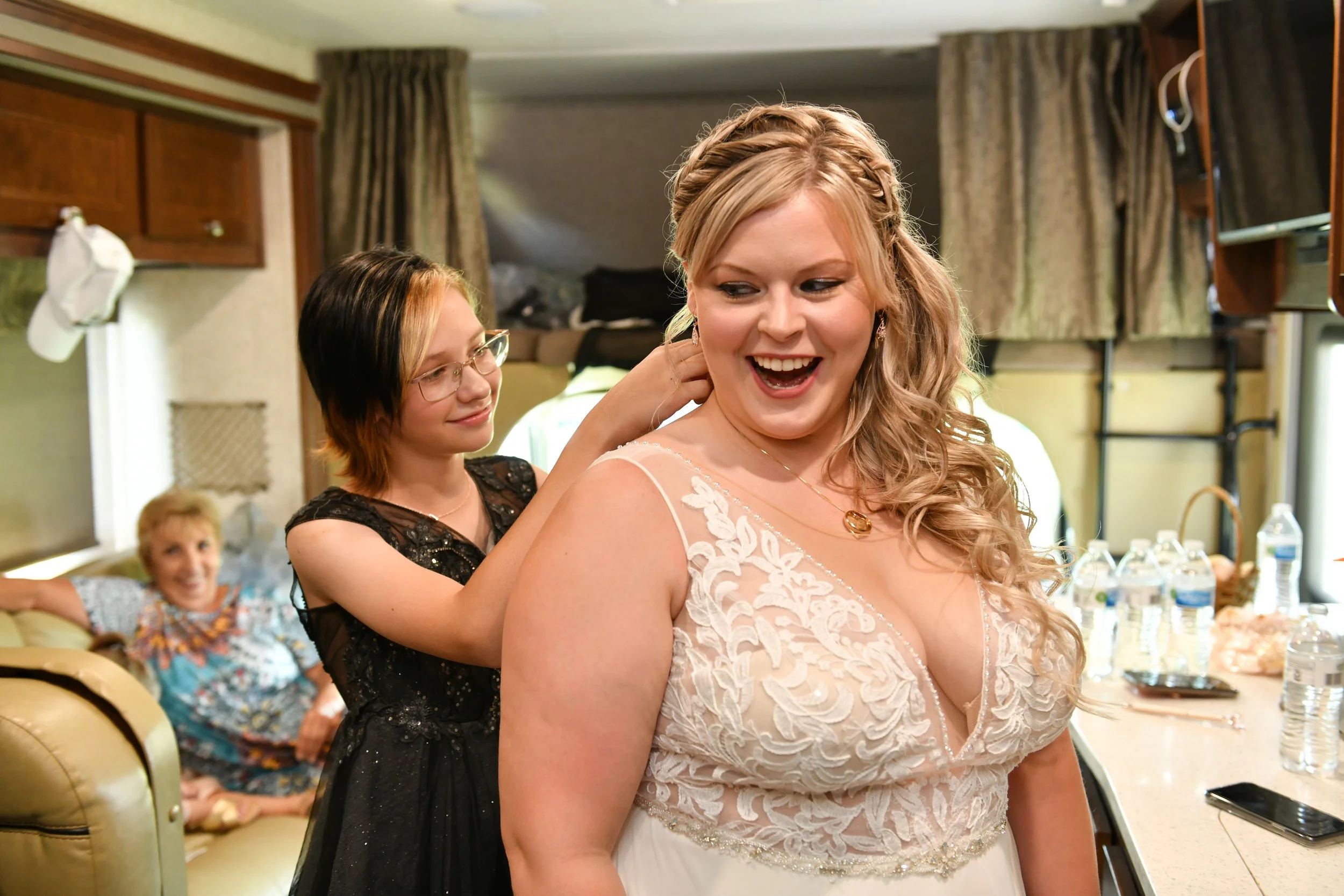 A woman in a wedding dress smiling as another woman helps her with her dress, while a third person is in the background smiling on a couch.
