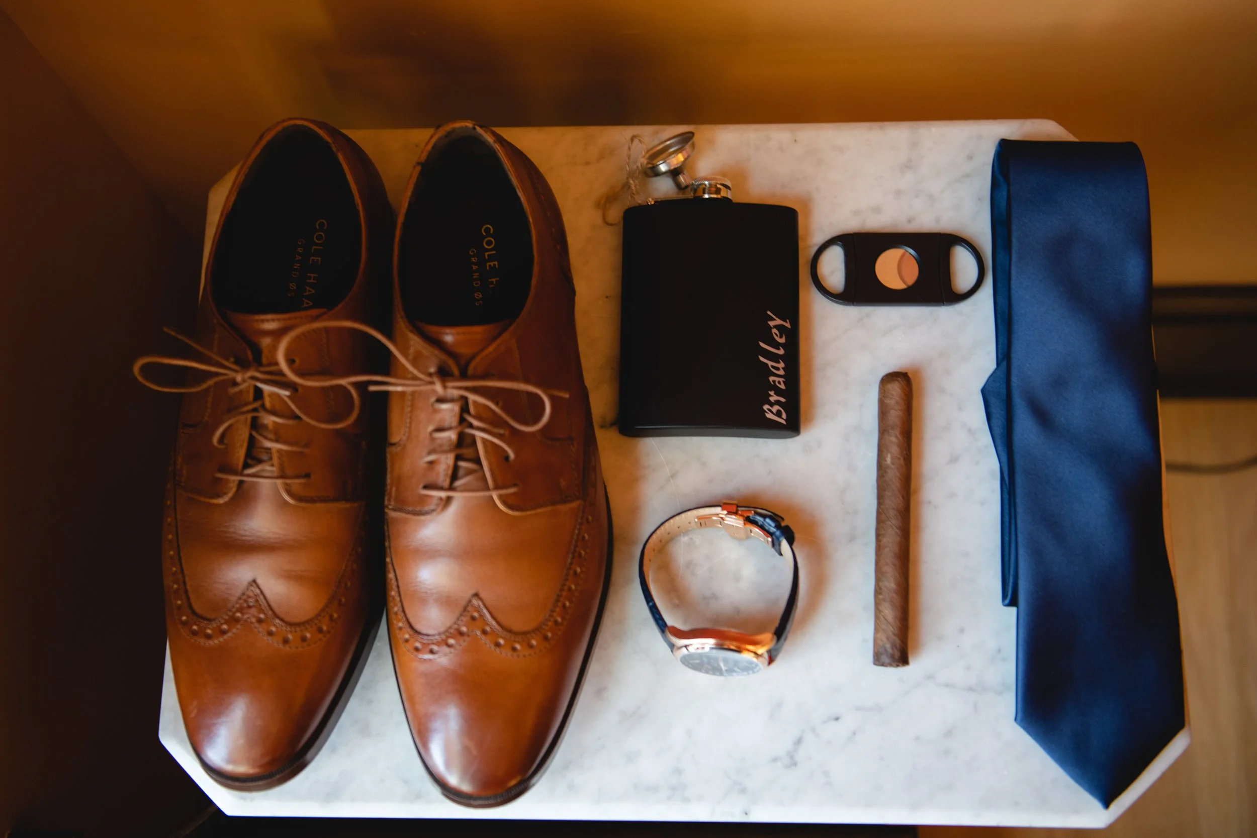Brown dress shoes, black wallet with "Bradley" written on it, black keychain, cigar, watch, brown cigar, blue tie, black headphone case, and black belt buckle on a white marble surface.