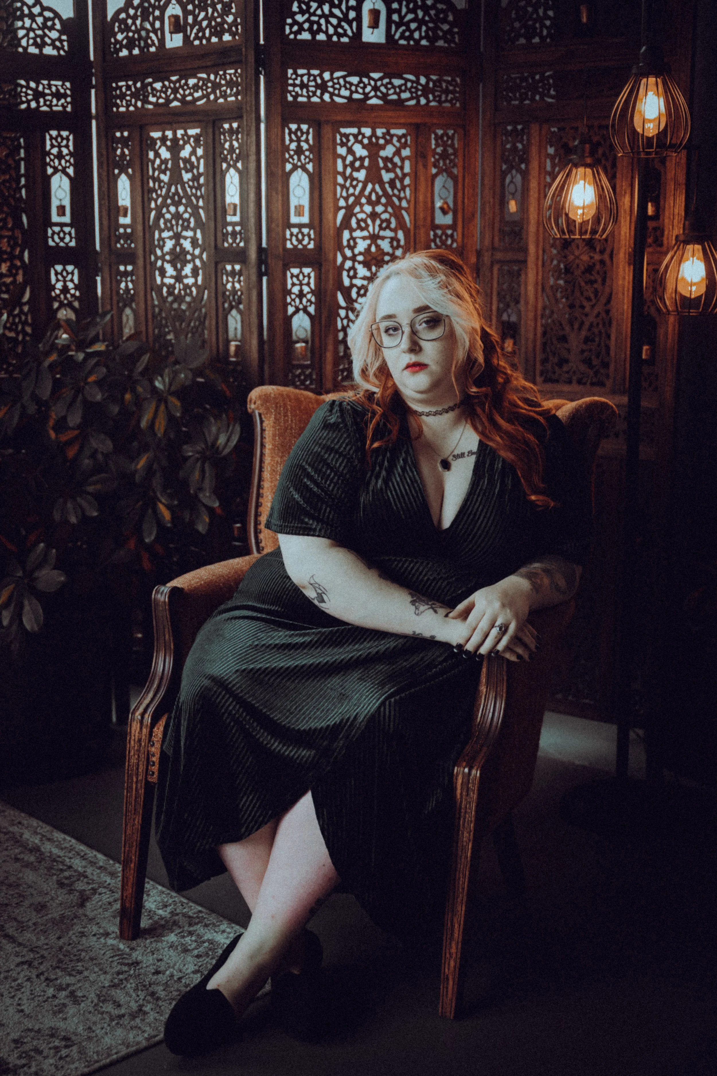 A woman with red hair and glasses sitting on a vintage armchair in a dimly lit room with ornate wooden screens and warm lighting fixtures.