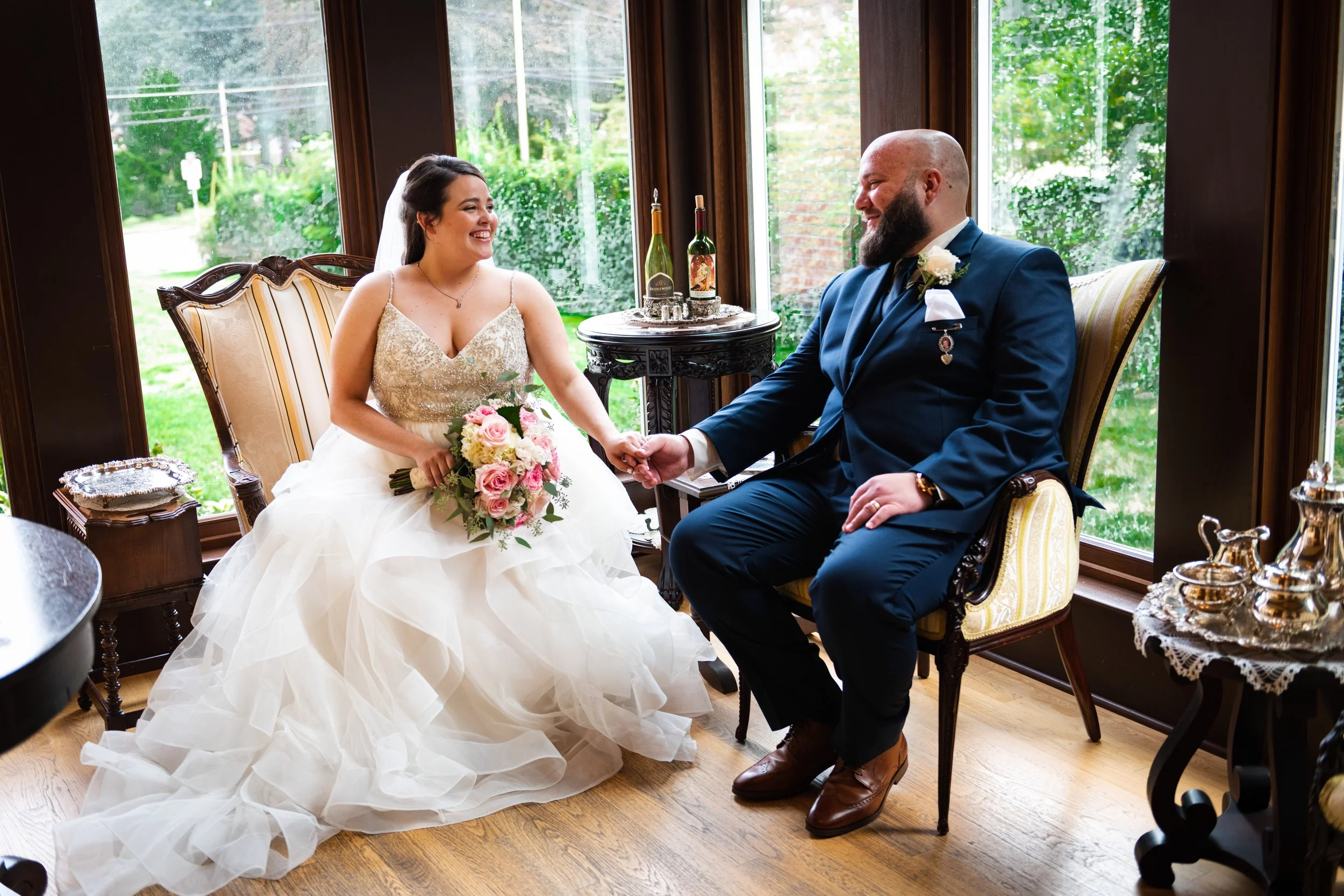 A bride and groom sit indoors. The bride wears a white wedding gown and holds a bouquet of pink roses. The groom is in a dark blue suit with medals and a white boutonniere. They are holding hands, smiling at each other, in front of large windows with
