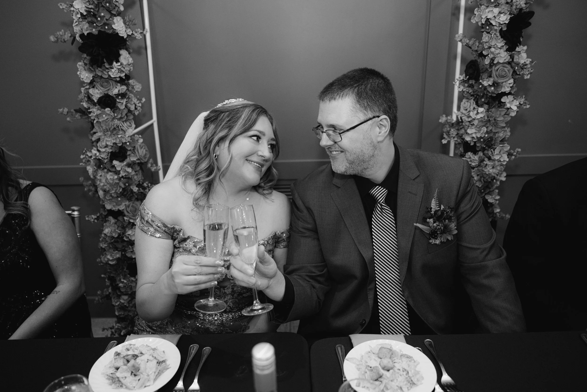 Couple at wedding reception toasting with champagne, woman wearing off-shoulder dress and veil, man in suit with boutonniere, decorated floral backdrop behind them, food on table in front.