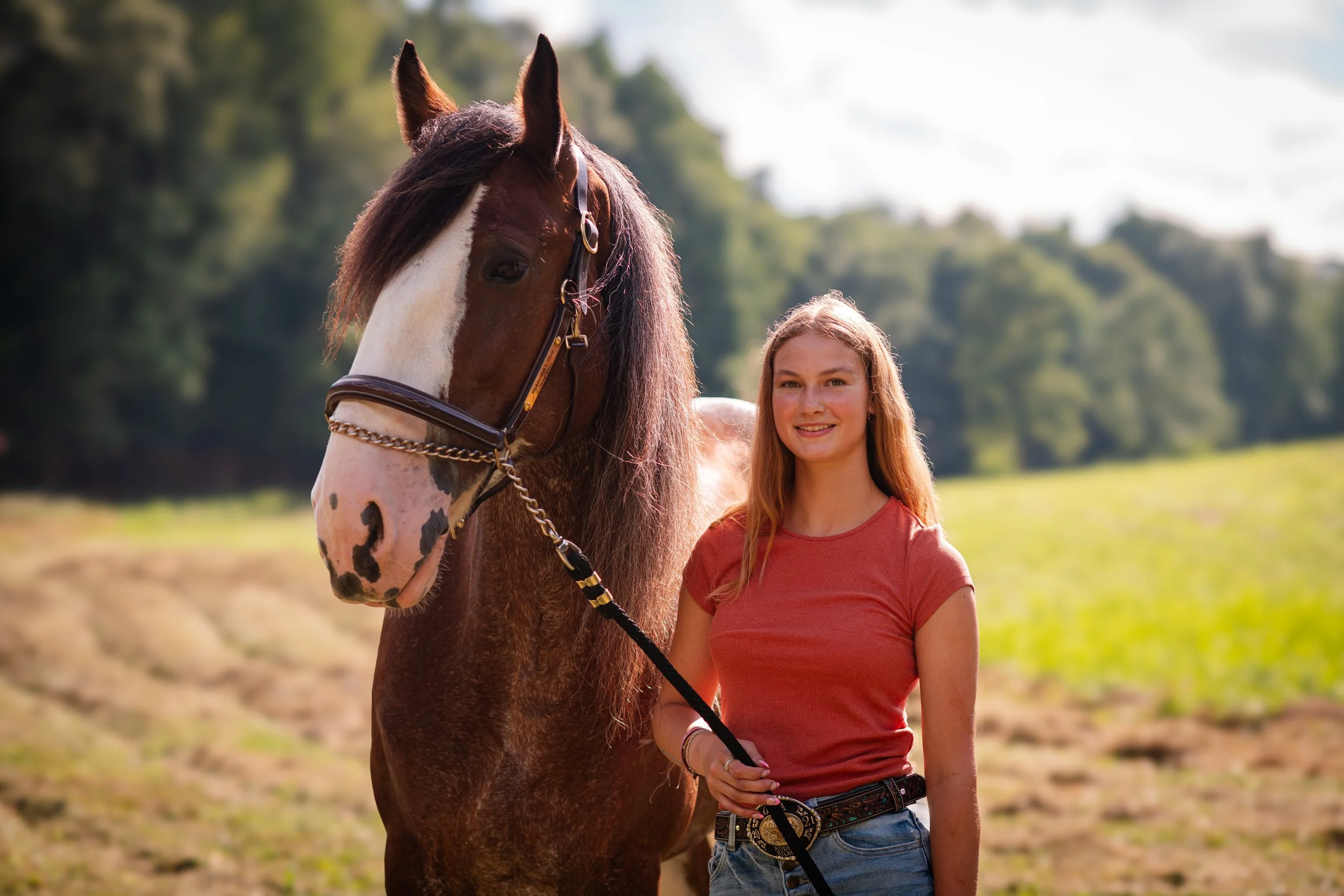 A young woman with long red hair wearing a pink t-shirt and blue jeans standing next to a large brown and white spotted horse outdoors in a field with green trees in the background.