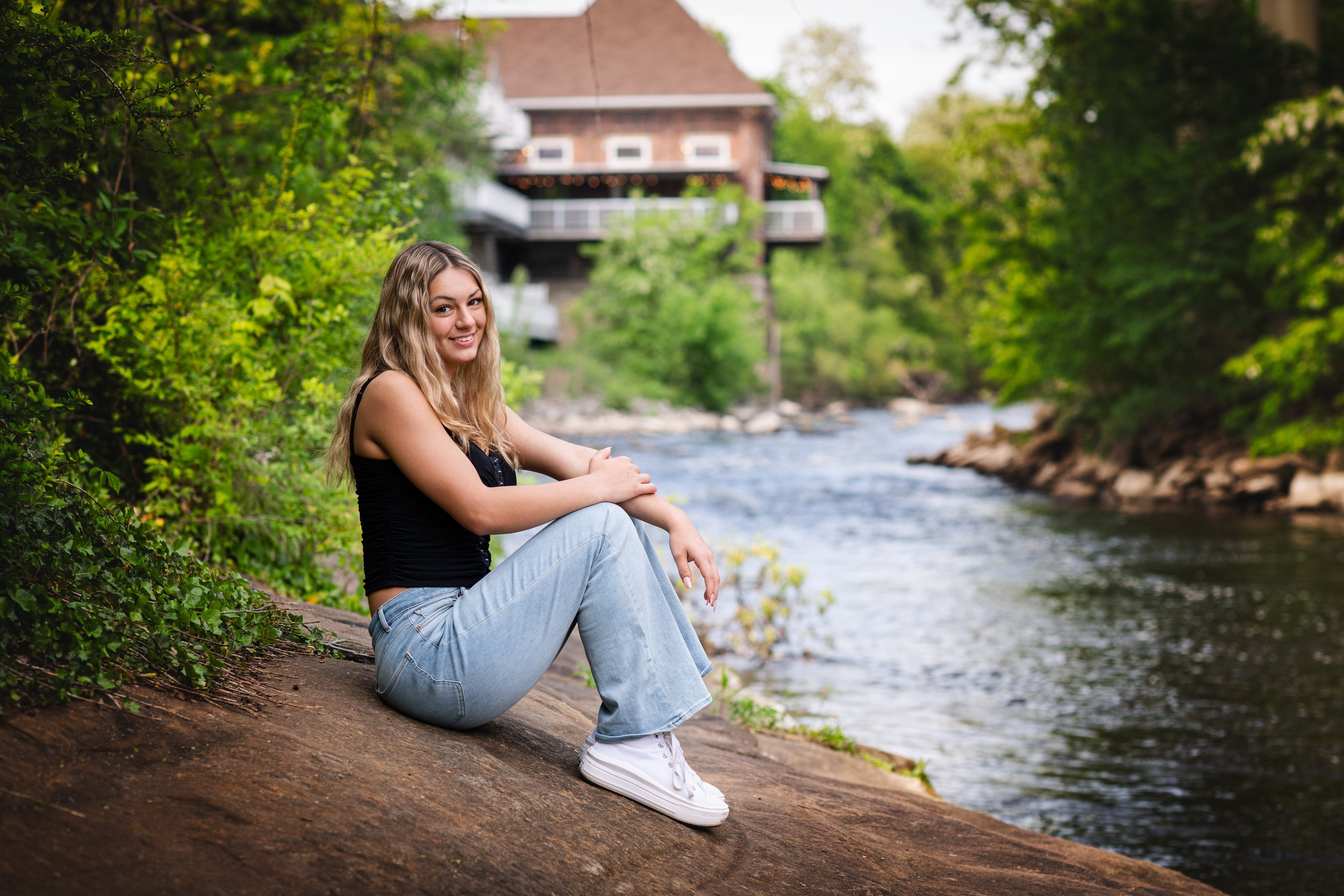 Young woman with long, wavy blonde hair, wearing a black tank top, light blue jeans, and white sneakers, sitting on a rock beside a river with green trees and a house in the background.