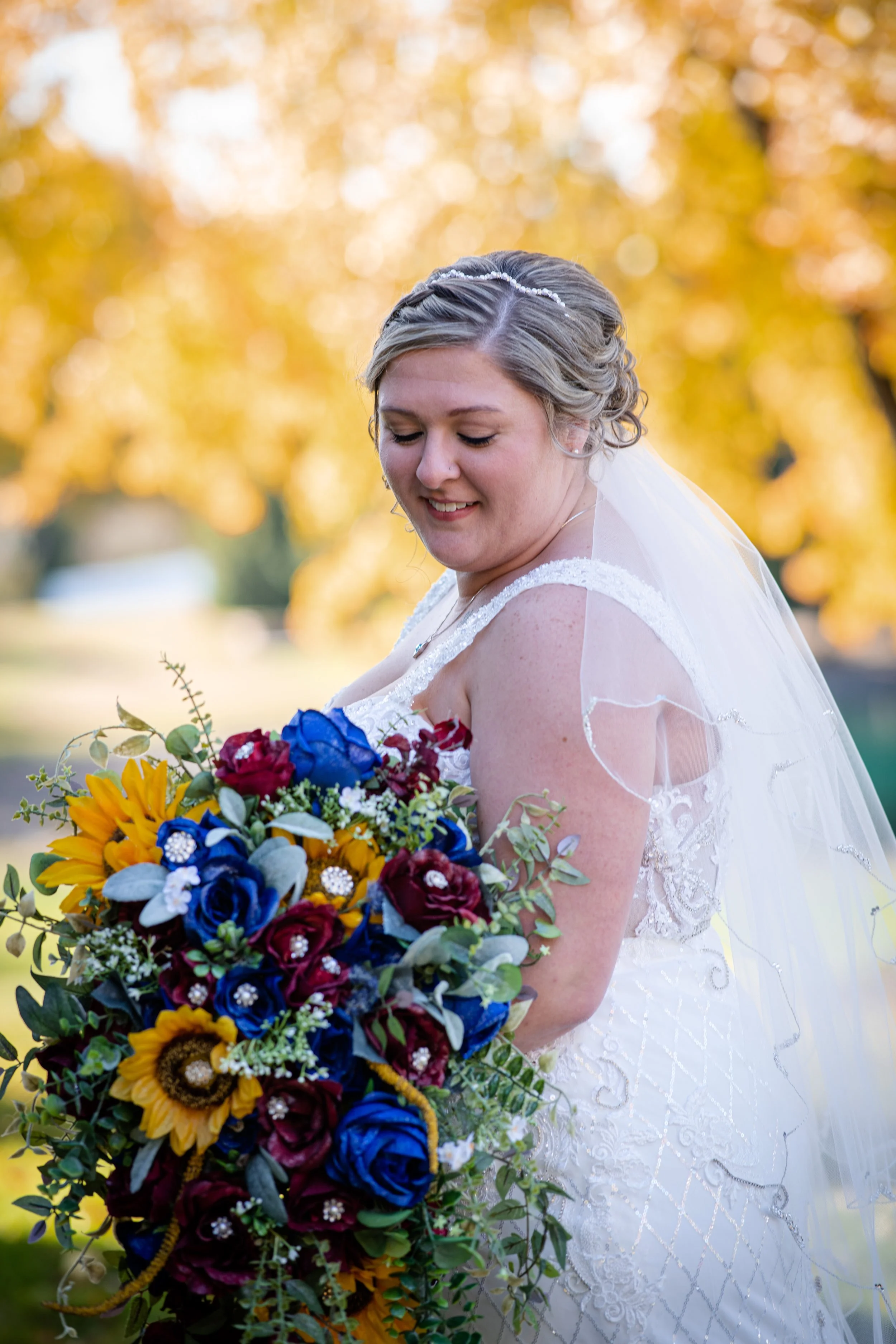 A bride wearing a white wedding dress and veil, standing outdoors with yellow autumn leaves in the background, holding a large colorful bouquet of sunflowers, red and blue roses, and greenery.