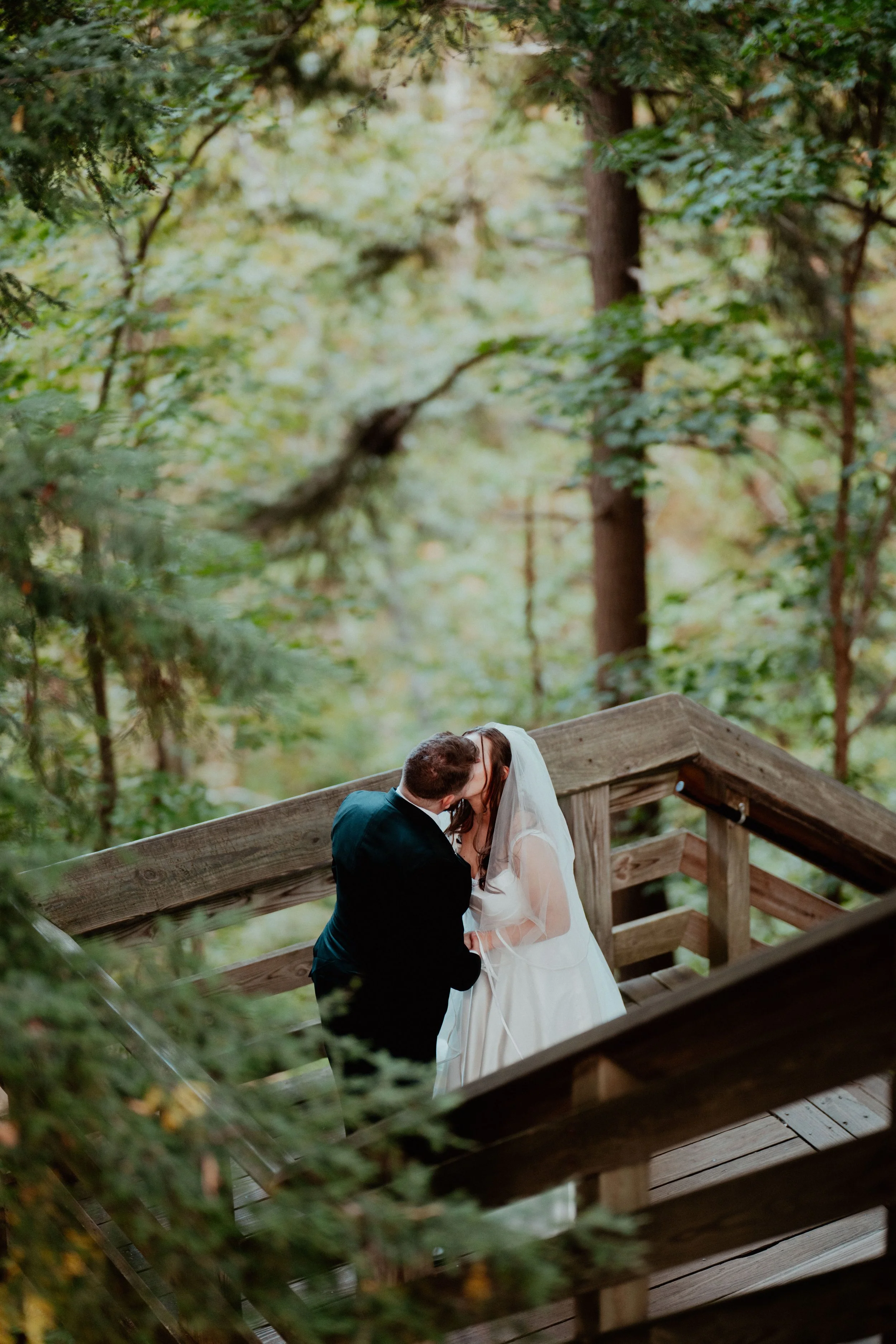A couple sharing a kiss on a wooden staircase surrounded by trees and greenery in a forest setting.