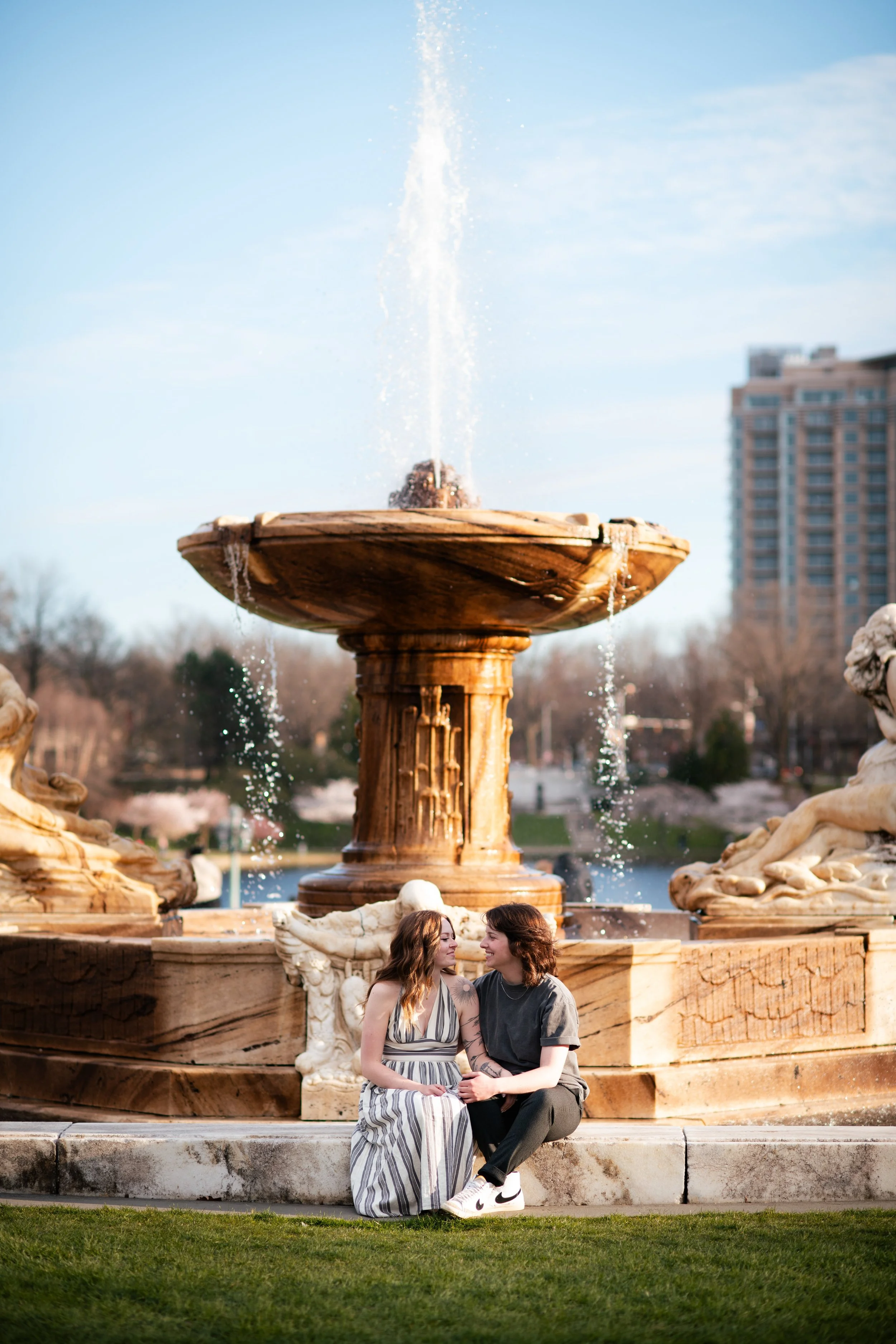 Two women sitting next to each other in front of a large decorative fountain with water spouting from the top, in a park.