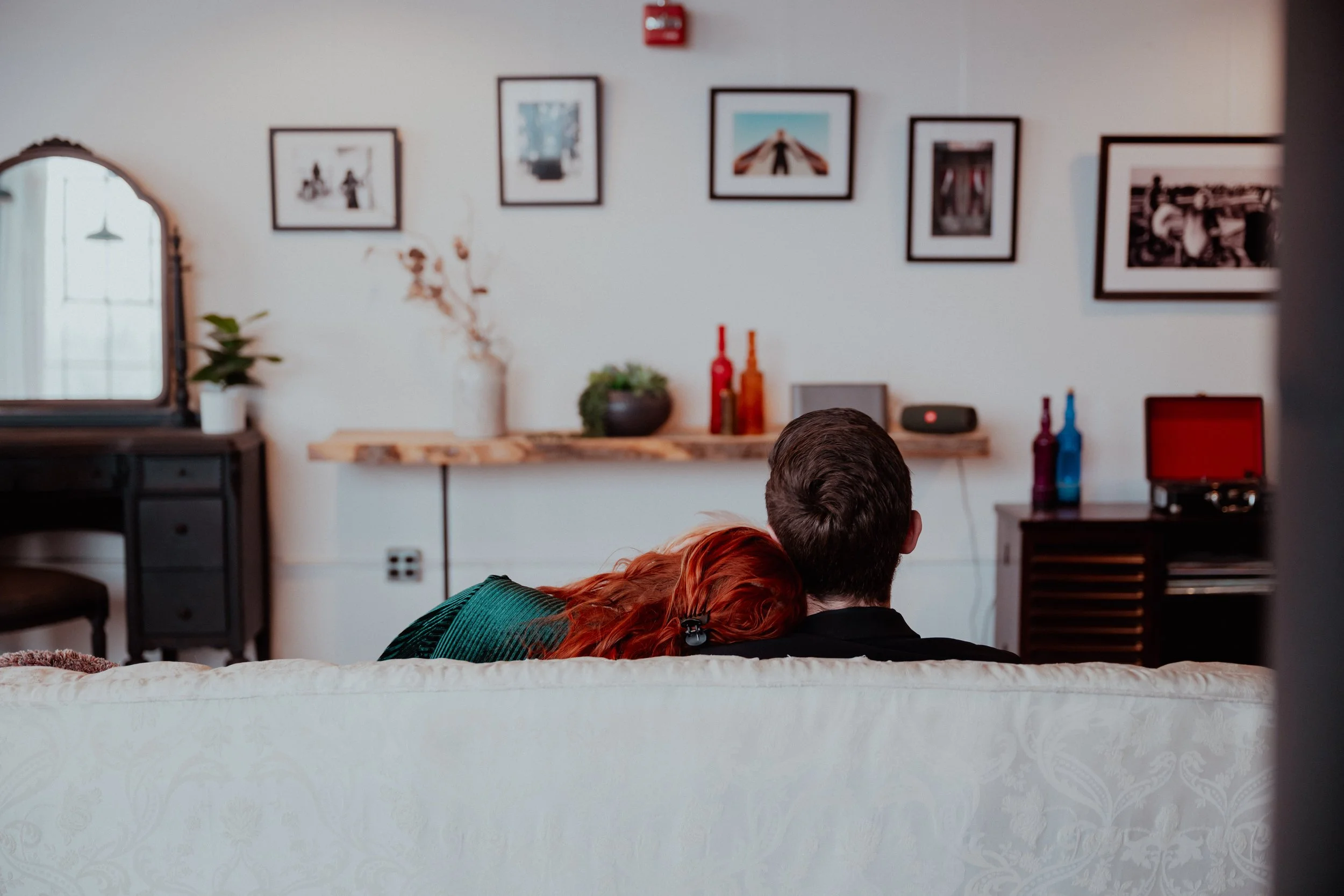 A couple sitting on a white sofa in a living room, with the woman resting her head on the man's shoulder. The room has a wall with framed photos, a mirror, and decorative bottles on a shelf.